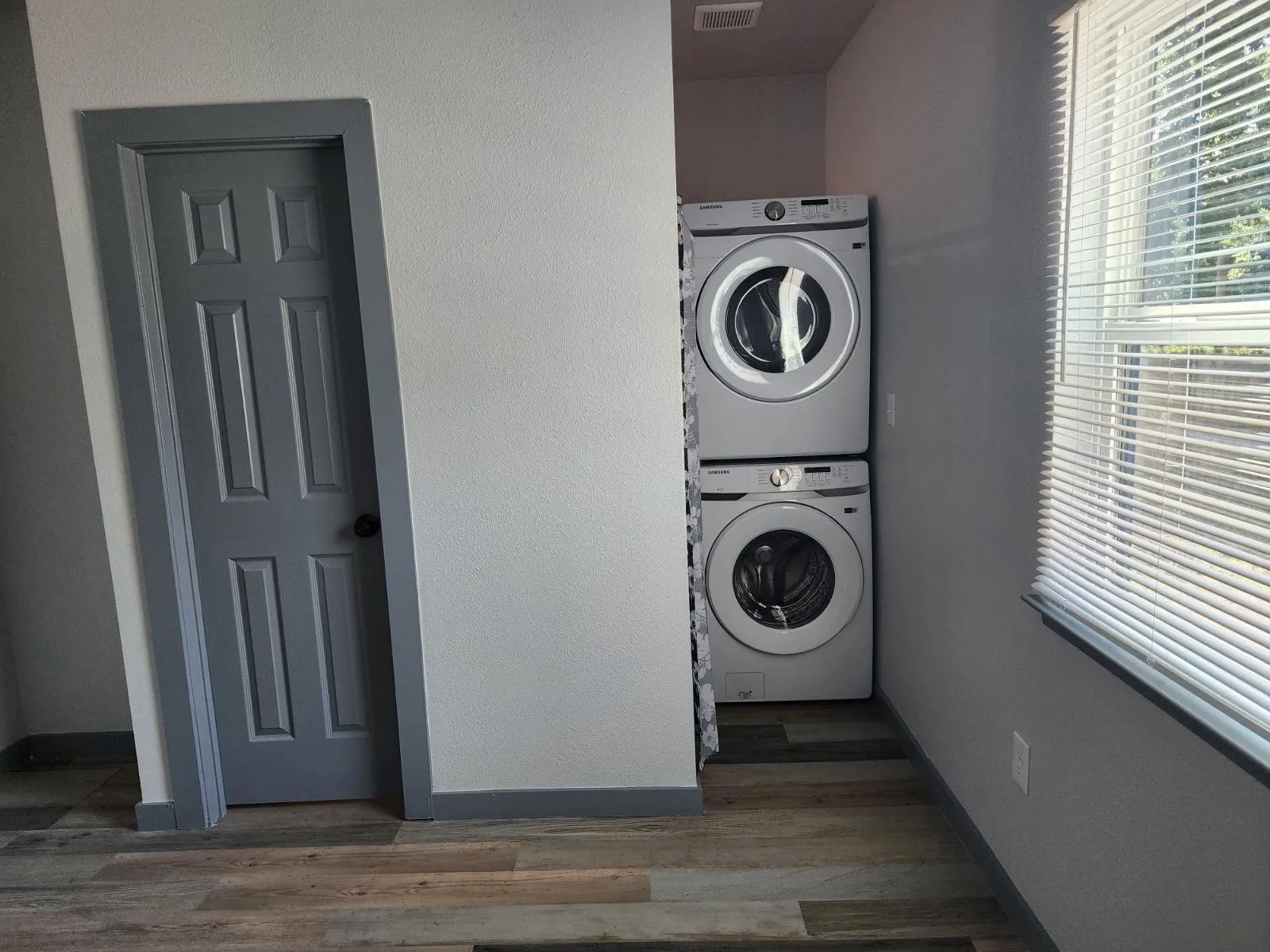 Stacked laundry appliances, wood-finish flooring, and a window with horizontal blinds