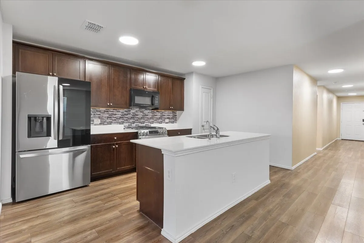 Kitchen featuring wood-finish flooring, recessed lighting, a stainless steel refrigerator, dark wood cabinetry, and a white countertop island with an integrated sink