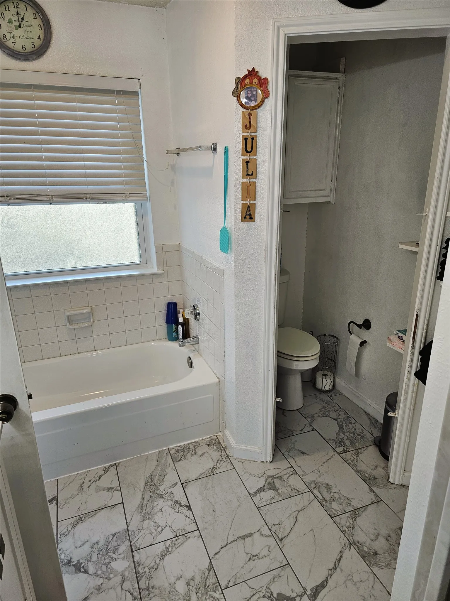 White ceramic tile tub with a window, white tile wainscoting, and large format marble-look floor tiles