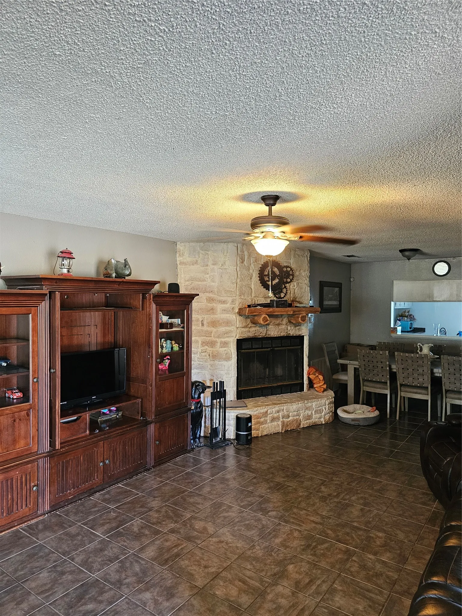 Stone-clad fireplace with a wood mantel, ceiling fan with integrated lighting, and ceramic tile flooring