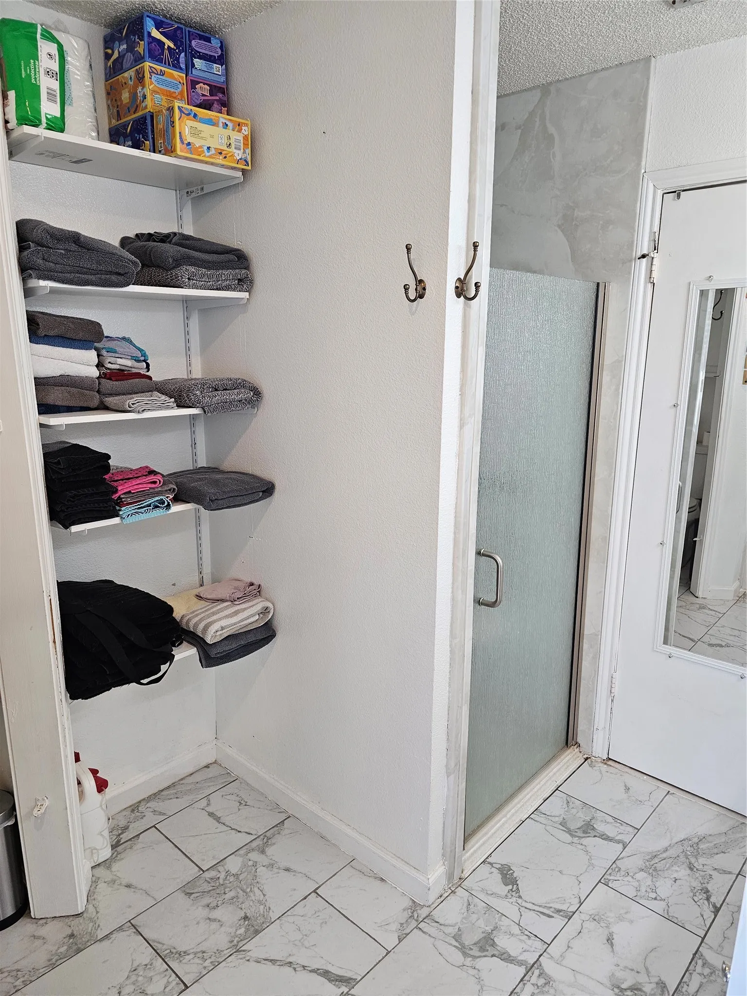 Shower enclosure with a frosted glass door, wall-mounted coat hooks, and white tile flooring with gray veining