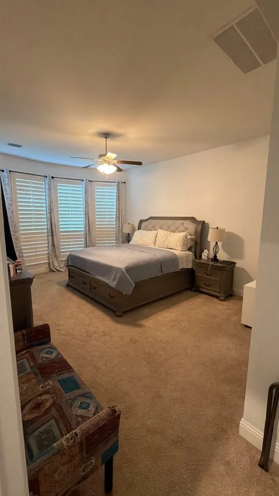 Carpeted bedroom featuring a ceiling fan with integrated lighting, multiple windows with plantation shutters, and a recessed ceiling speaker