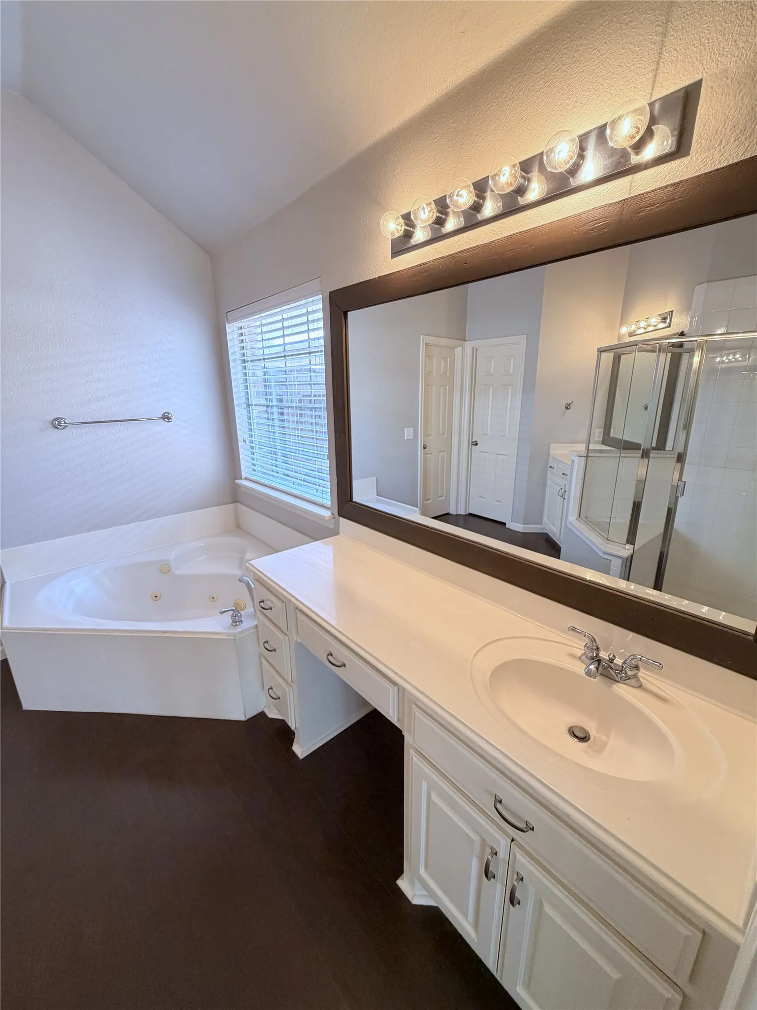 Bathroom featuring a corner jetted soaking tub, dark wood-finish flooring, white vanity with integrated sink, chrome faucet, and a large wall mirror with overhead lighting