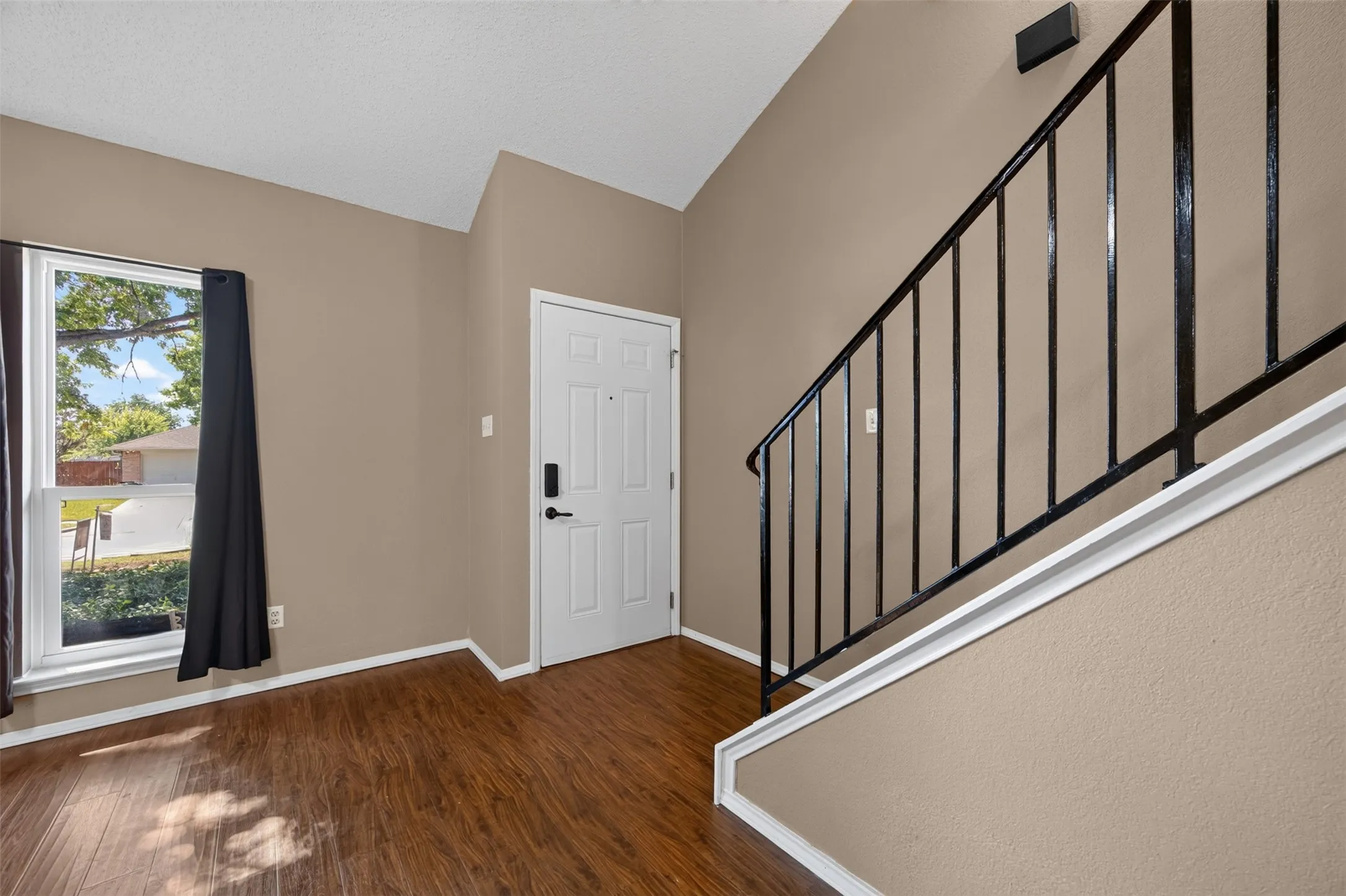 Vaulted ceiling, wood-finish flooring, white paneled entry door, black wrought iron railing, and a large window