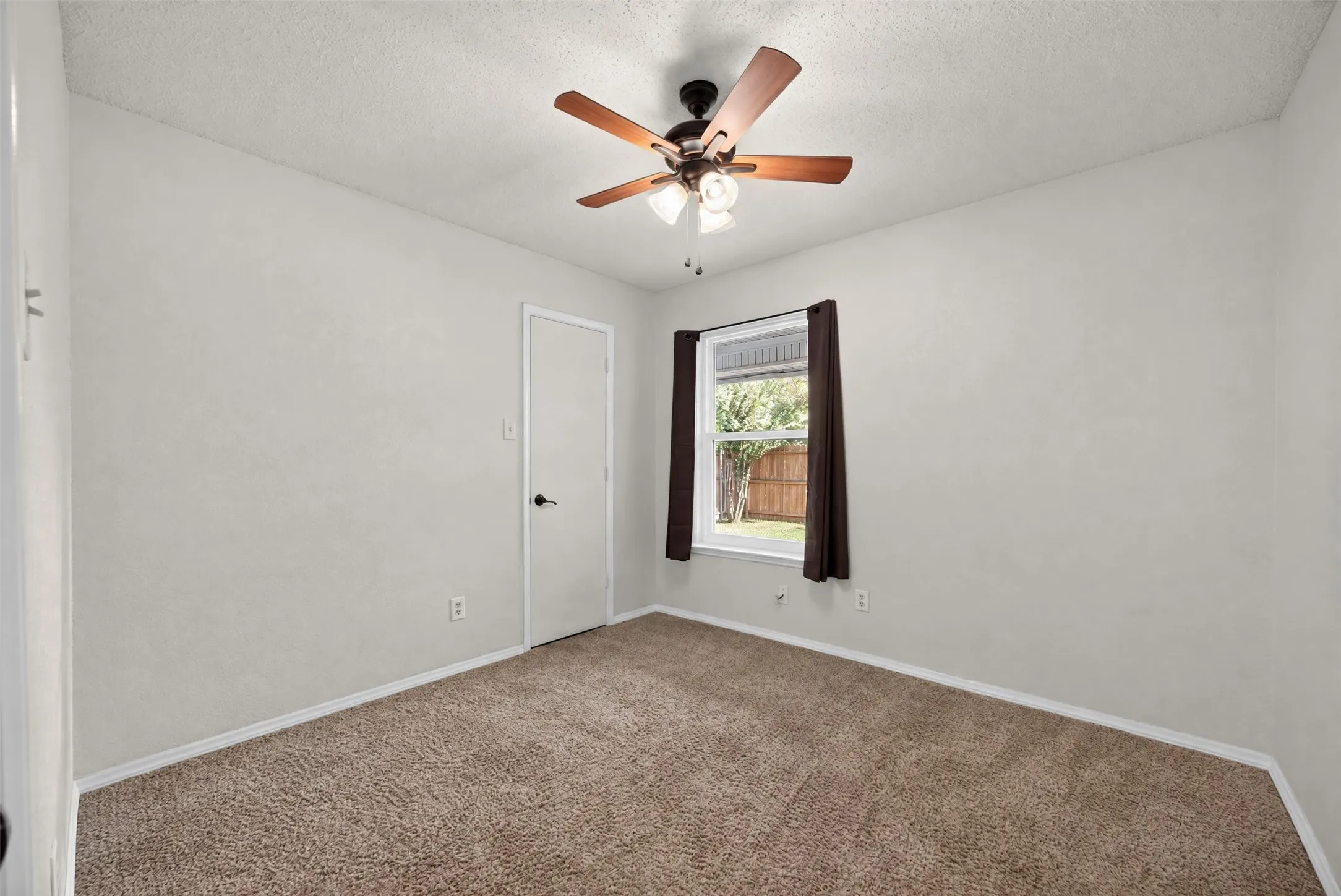 Neutral wall paint with textured finish, ceiling fan with integrated lighting, window with exterior yard visibility, and brown carpet flooring