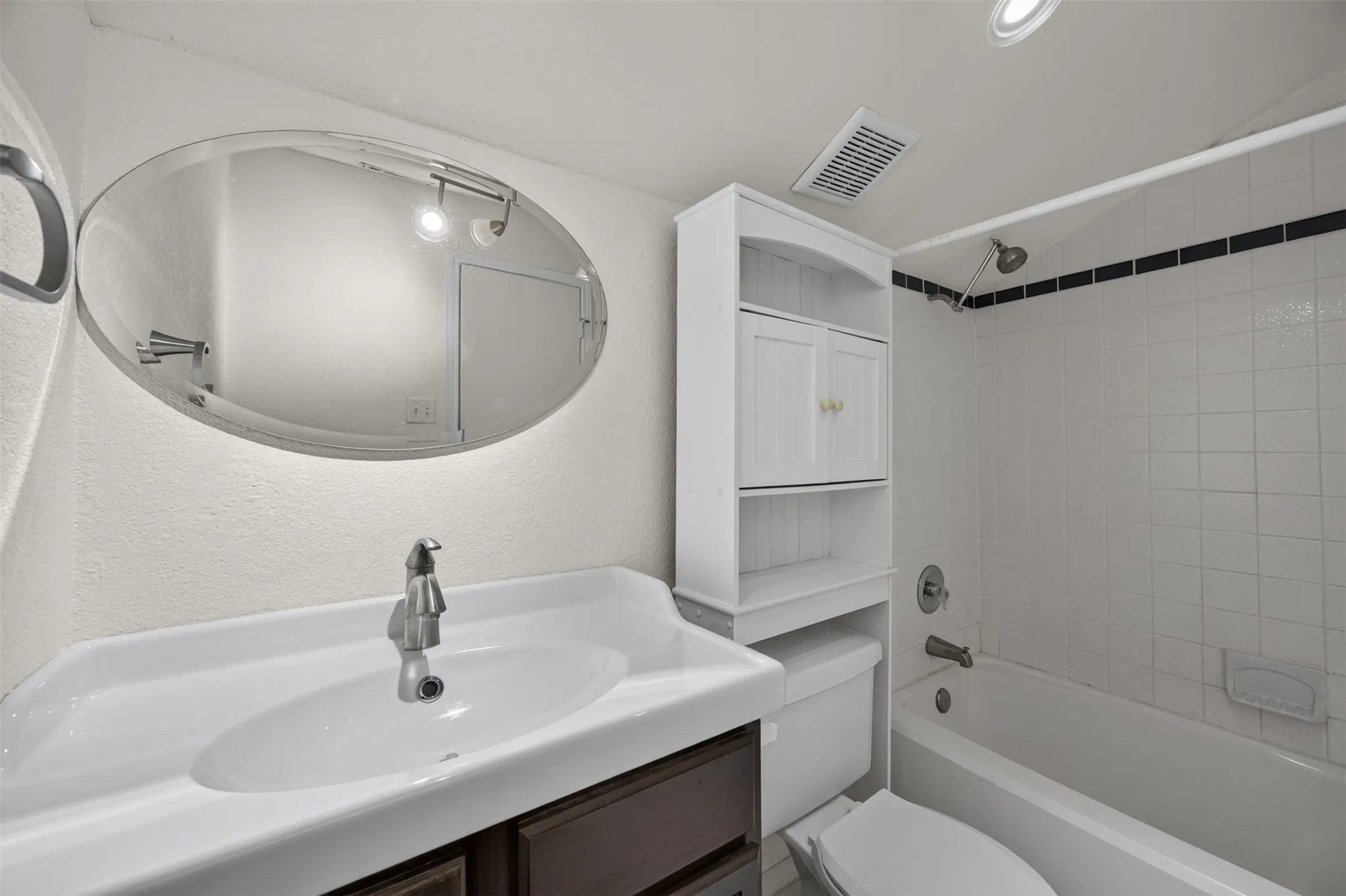 Bathroom featuring a white ceramic vanity sink, a wall-mounted oval mirror, a white built-in storage cabinet, a white bathtub with tile surround, and a chrome shower fixture