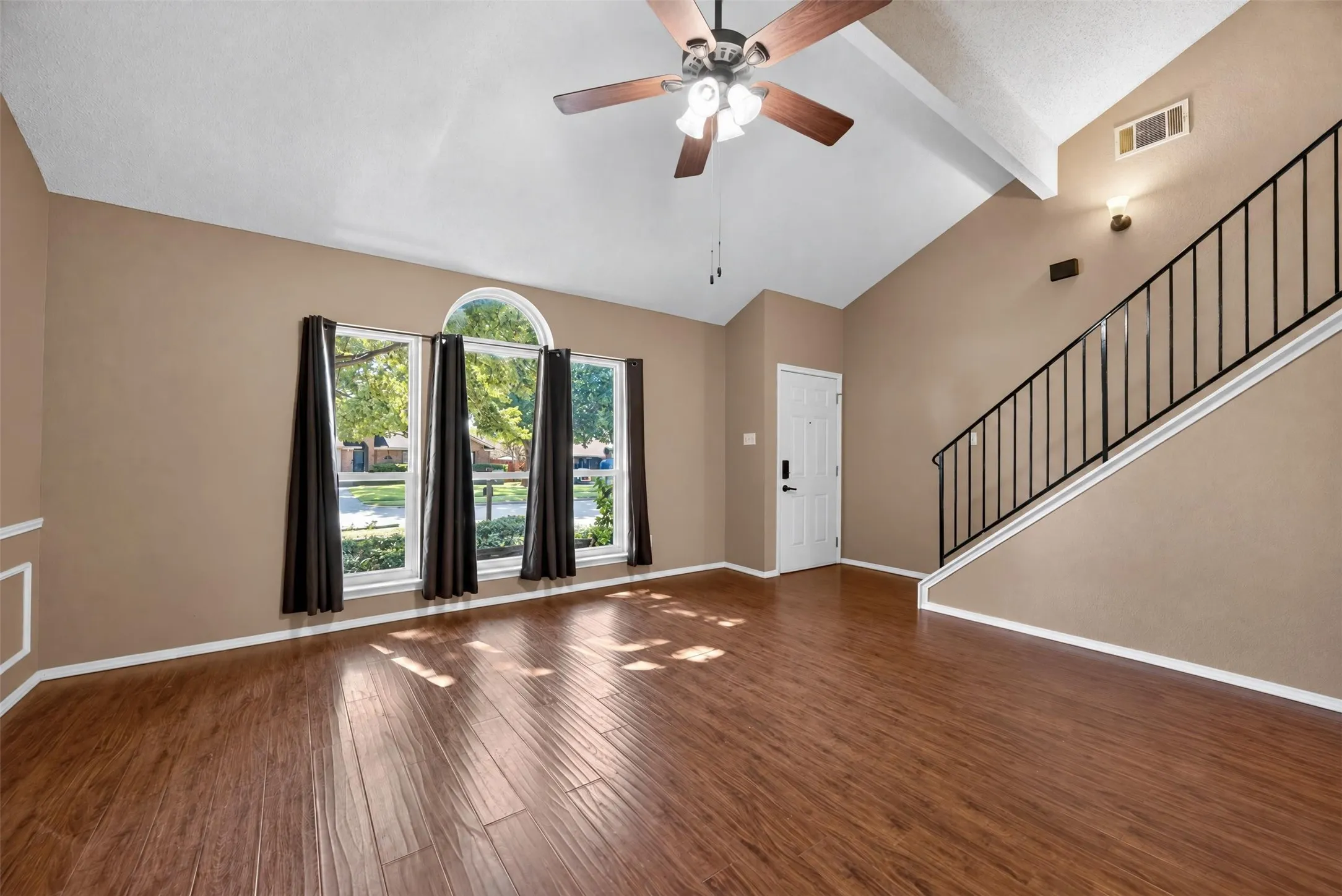 Vaulted ceiling with a ceiling fan, wood-finish flooring, staircase with a black metal railing, prominent window featuring an arched transom, and a solid entry door