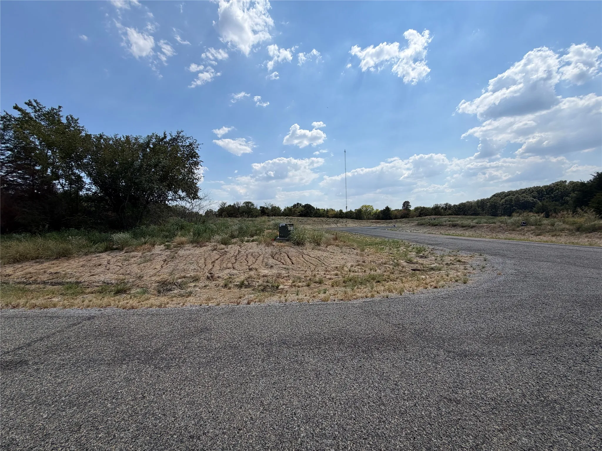 Paved road frontage with gravel shoulder, expansive clear sky, mature trees, and natural ground cover