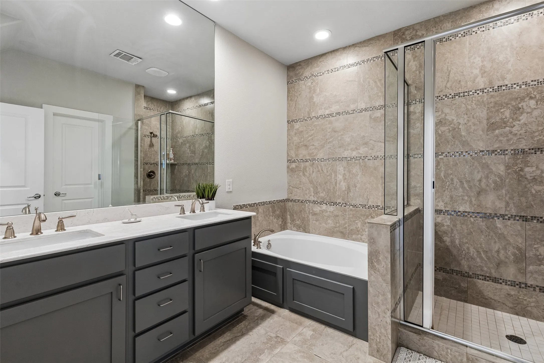 Dual vanity with individual sinks, white countertop, dark gray cabinetry, built-in bathtub, and a separate glass-enclosed shower with tiled walls