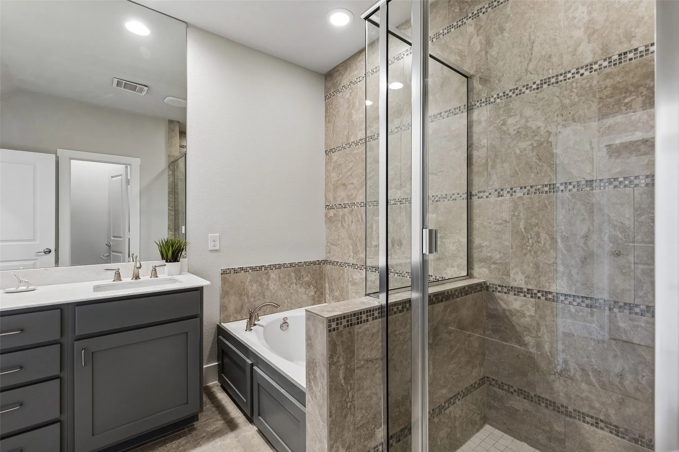 Vanity with undermount sink and brushed nickel fixtures, built-in bathtub, glass-enclosed shower, and tile-surround walls with mosaic accents