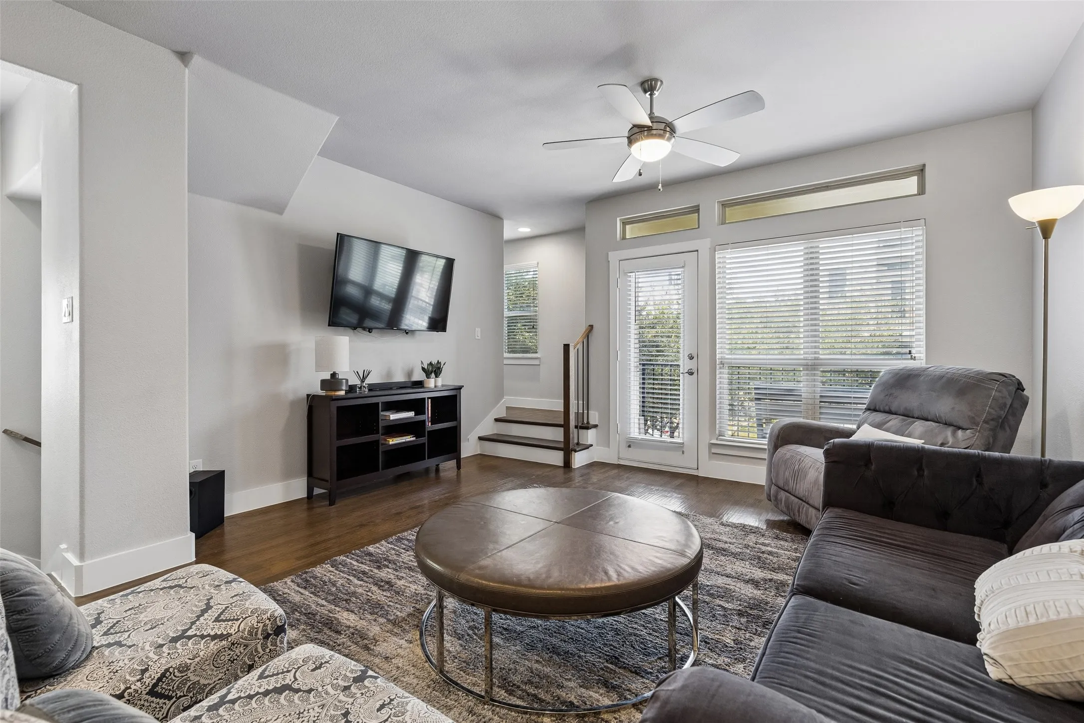 Hardwood-finish flooring, ceiling fan with integrated lighting, door with transom windows, white blinds, and interior staircase with dark wood treads