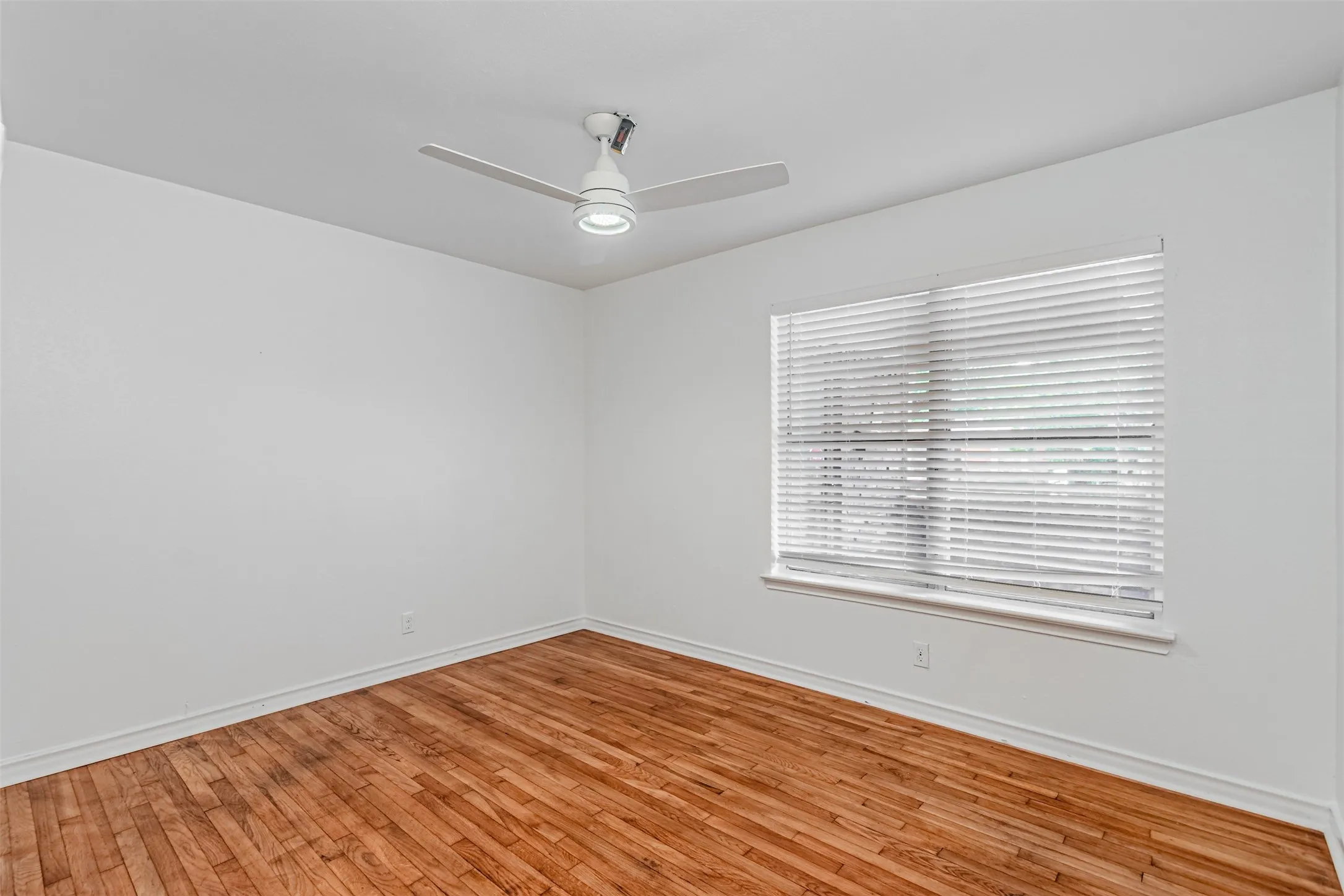 Wood-finish flooring with white baseboards, a window with blinds, a white ceiling fan with integrated lighting, and white painted walls