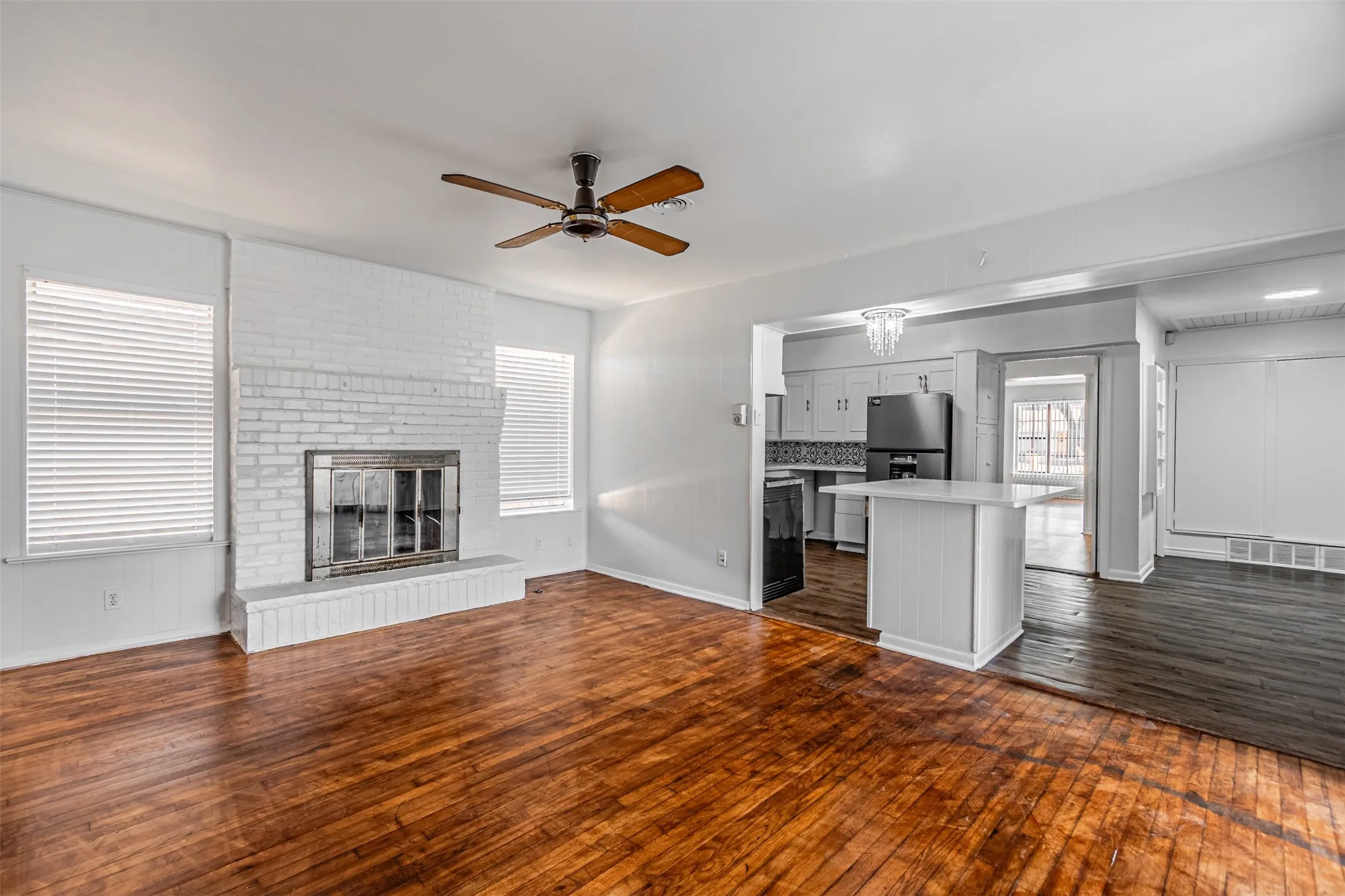 Wood-finish flooring, white painted brick fireplace with hearth, ceiling fan, and a kitchen pass-through with breakfast bar