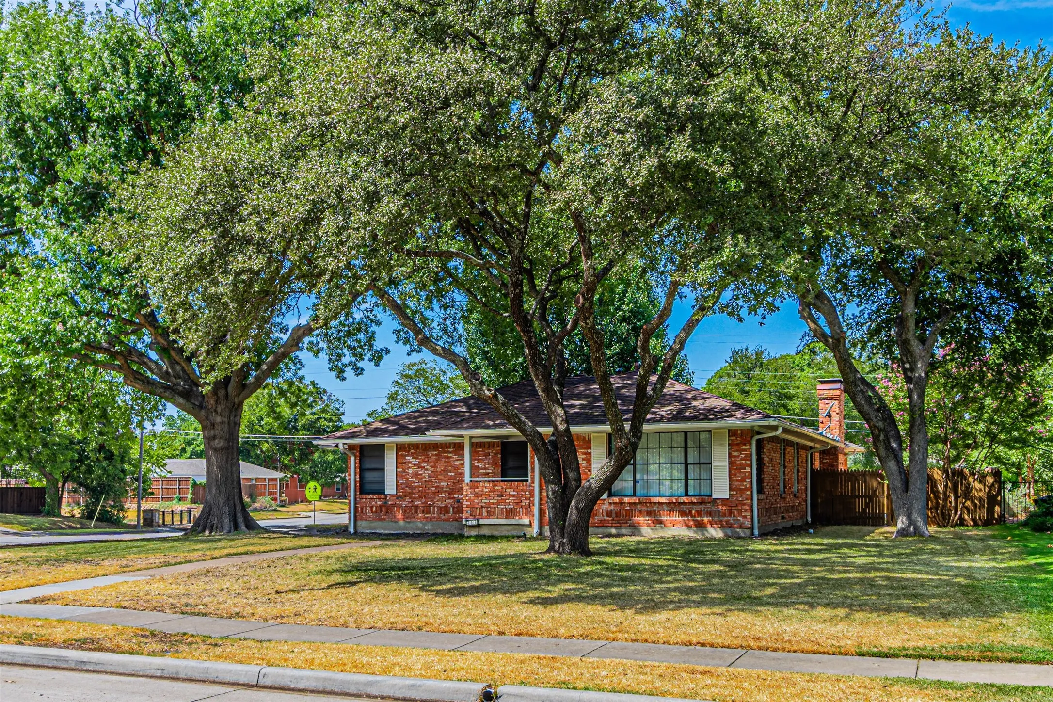 Brick facade residence featuring a shingled roof, mature trees, and a concrete sidewalk
