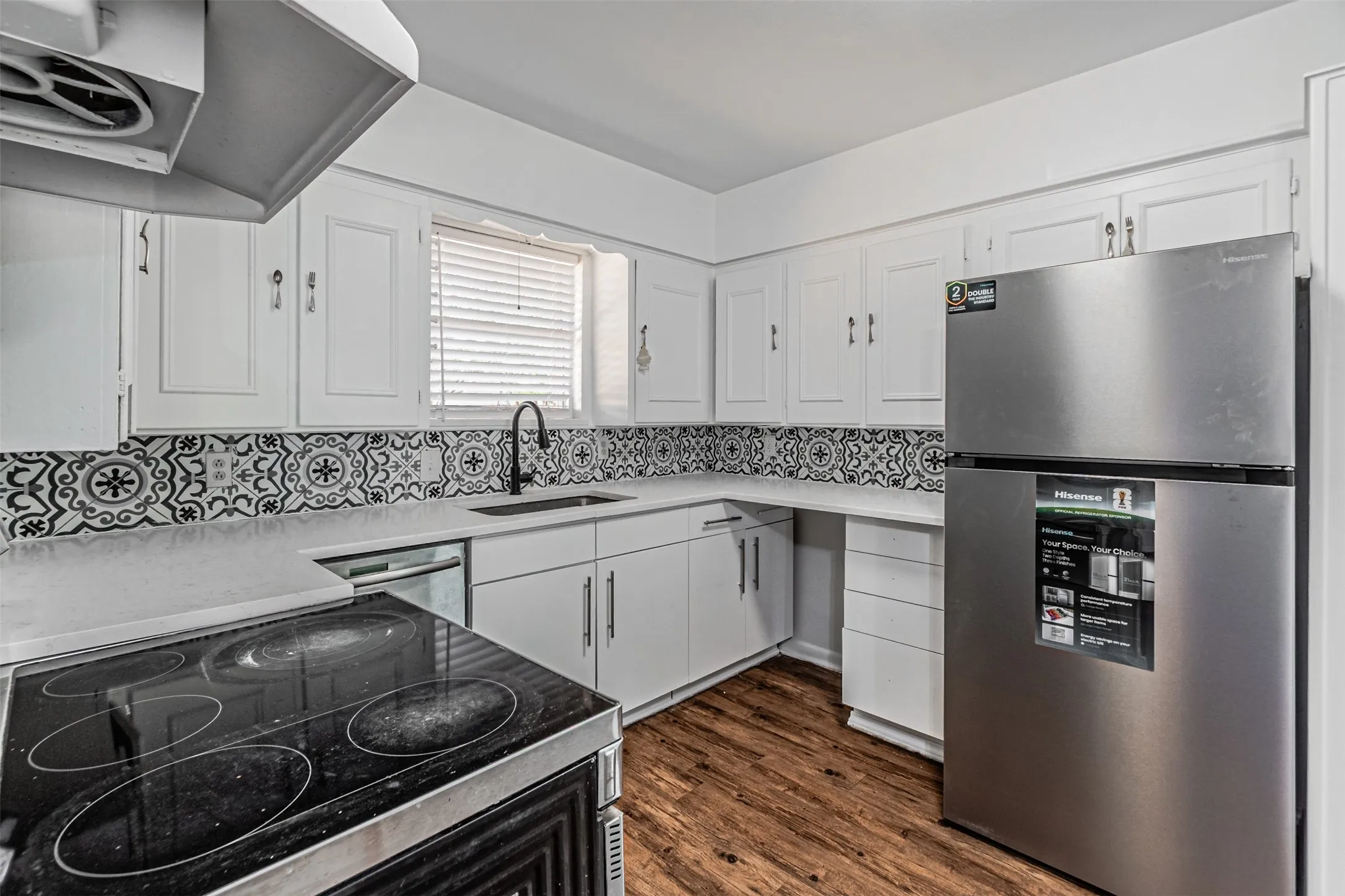White cabinetry, patterned backsplash, black-finish faucet, stainless steel refrigerator, and wood-finish flooring