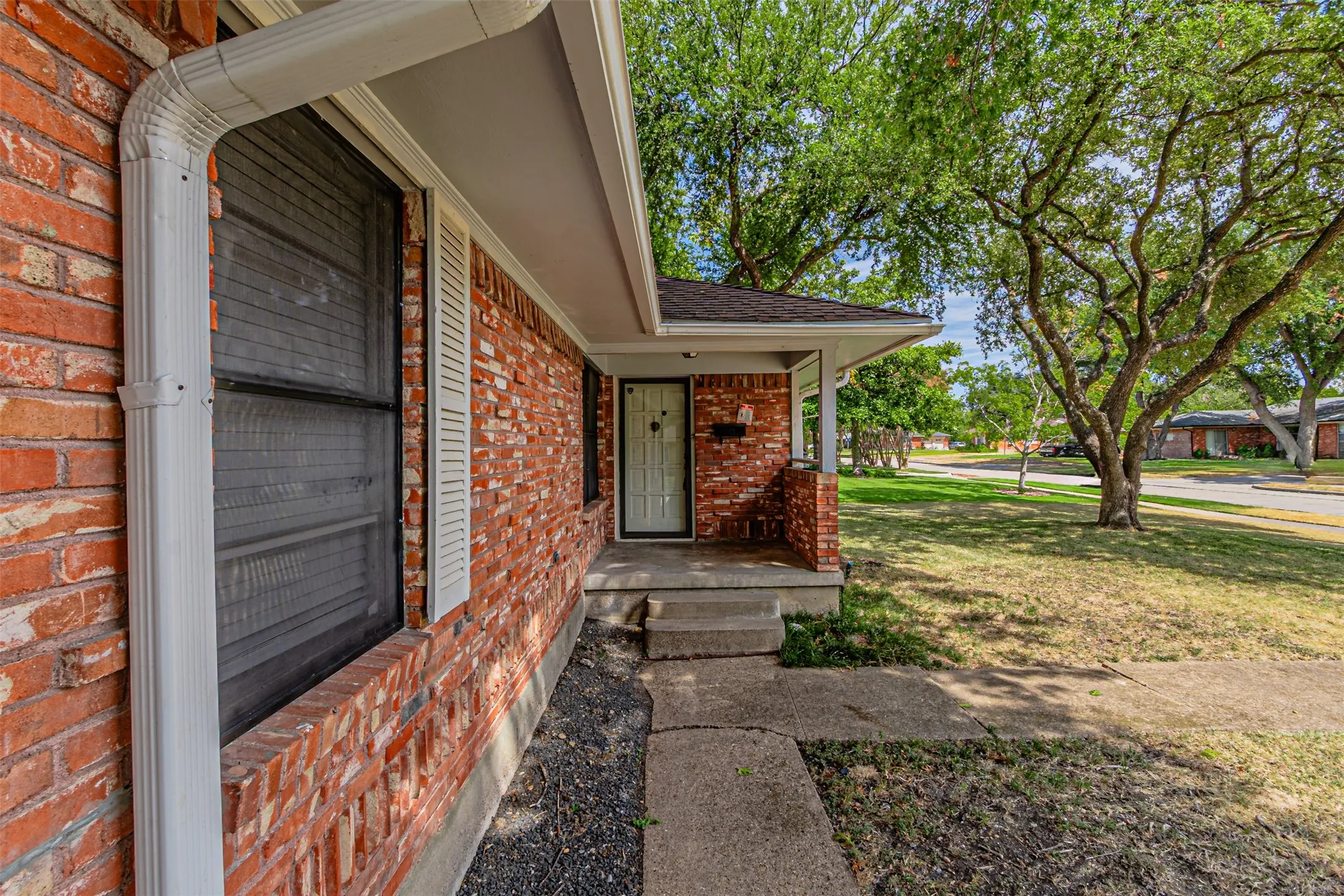 Brick exterior with white gutters and shutters, featuring a covered porch entry and mature trees