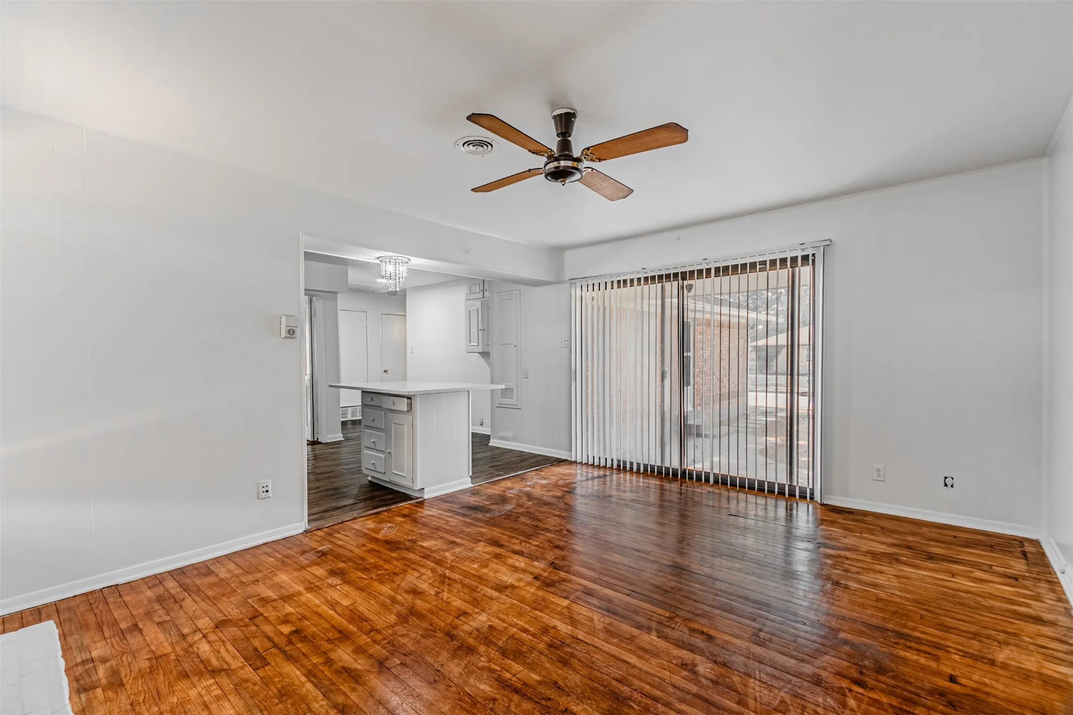 Wood-finish flooring throughout, ceiling fan with light fixture, sliding glass door with vertical blinds, and a pass-through opening to a kitchen with white cabinetry