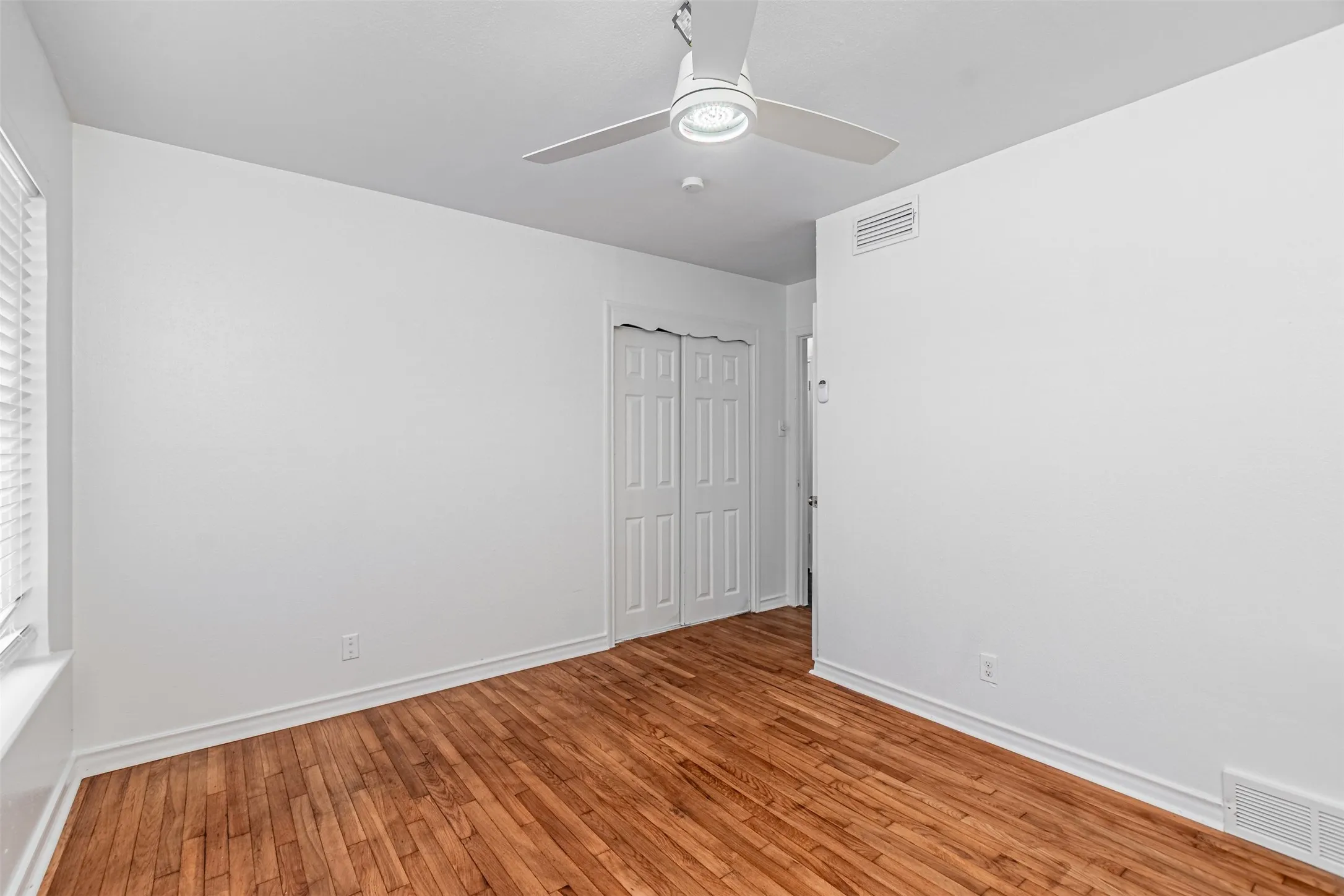 Wood-finish flooring, white walls, louvered closet doors, window with blinds, and a ceiling fan with integrated lighting