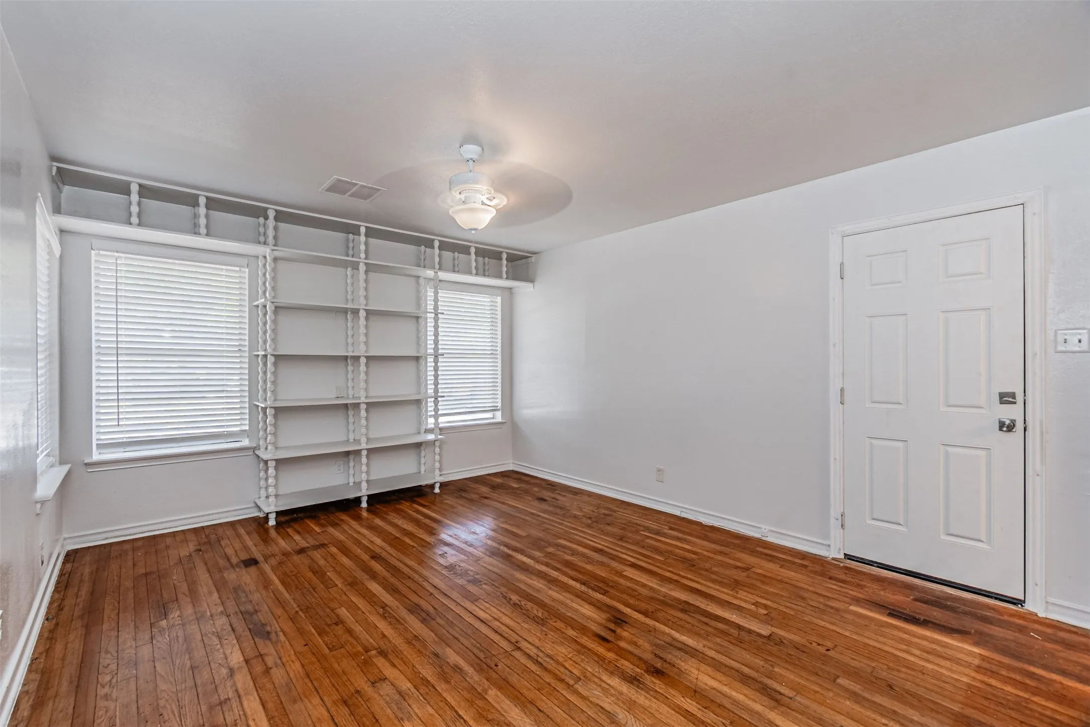 Hardwood flooring, two windows with blinds, built-in shelving, white paneled door, and ceiling light fixture