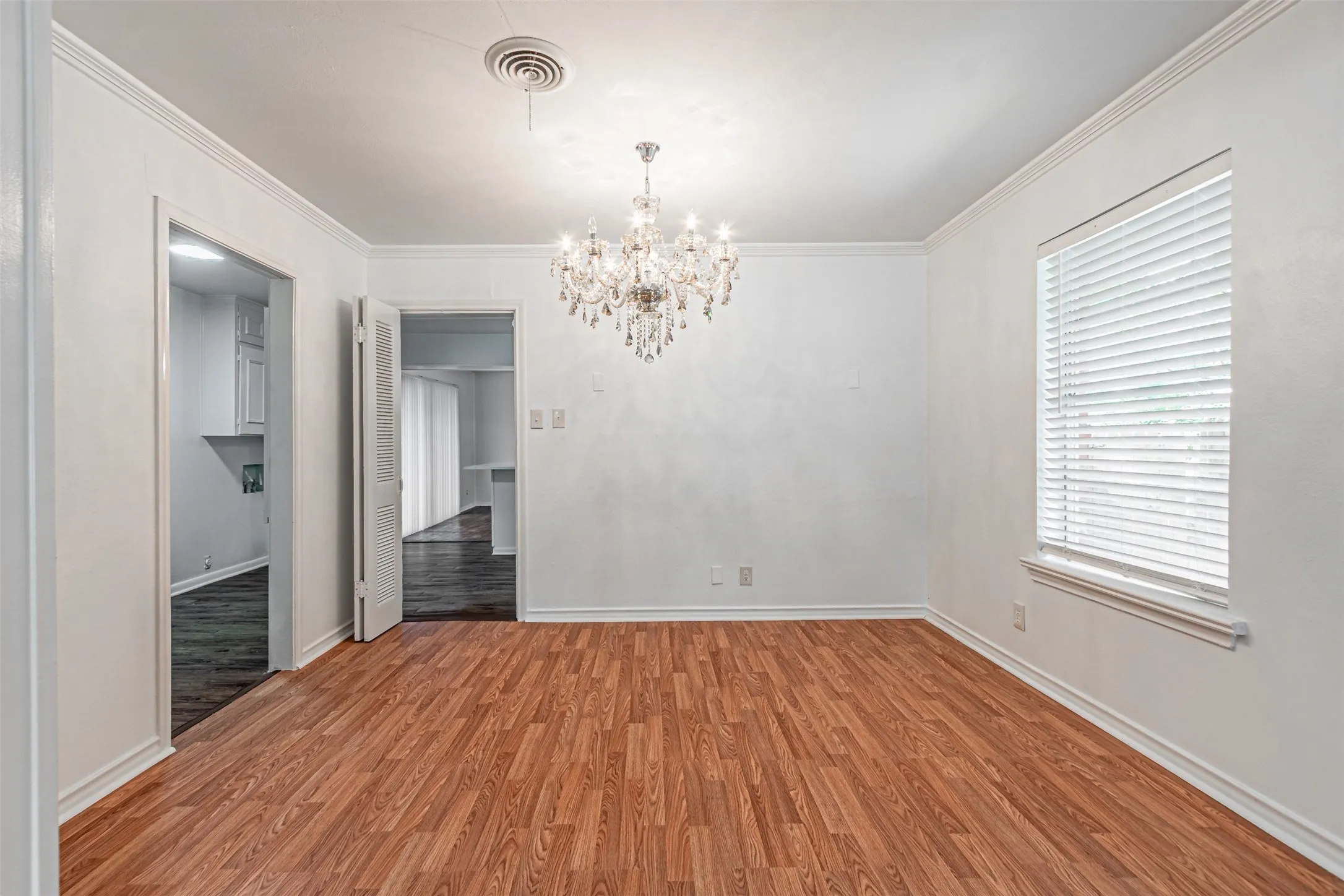 Wood-finish flooring, white walls, crown molding, a window with blinds, and a crystal chandelier