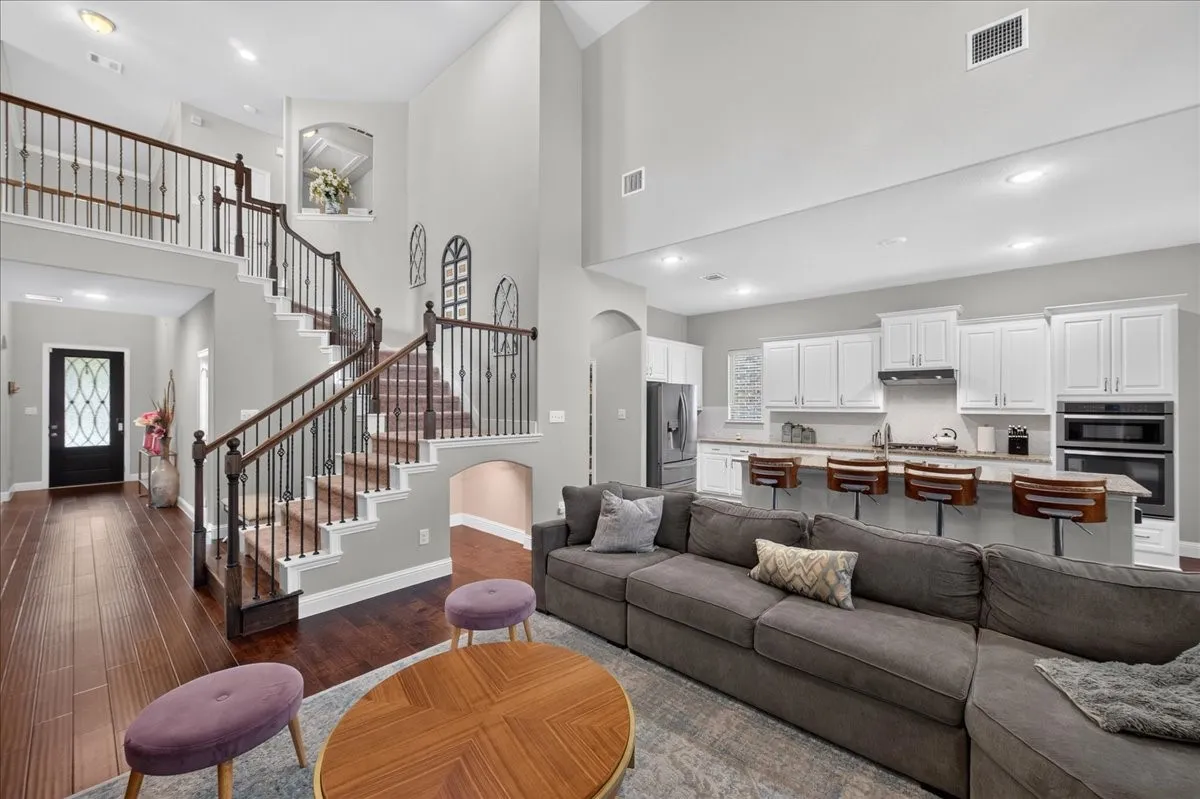 Two-story living area with high ceilings, wood-finish flooring, and an open-concept kitchen featuring white cabinetry and stainless steel appliances