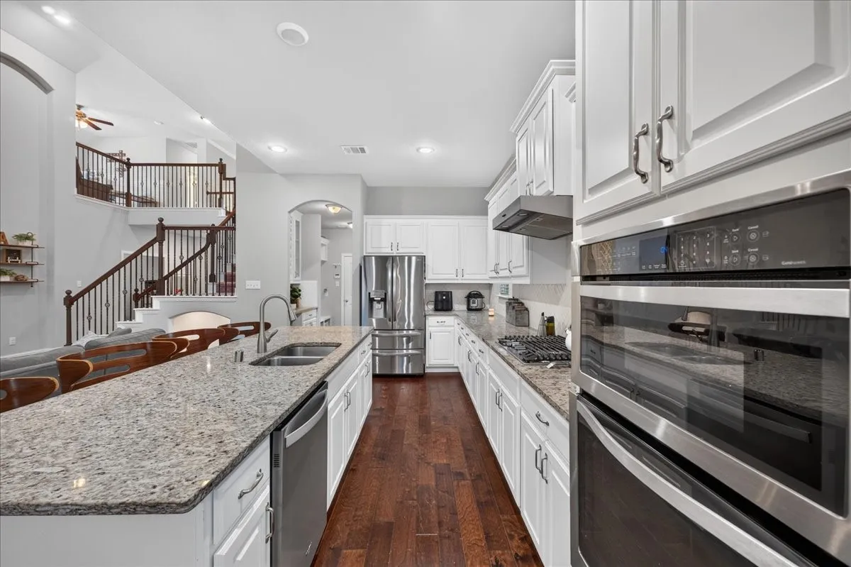 Spacious kitchen featuring white cabinetry, stainless steel appliances, granite countertops, an island with an undermount sink, and wood-finish flooring