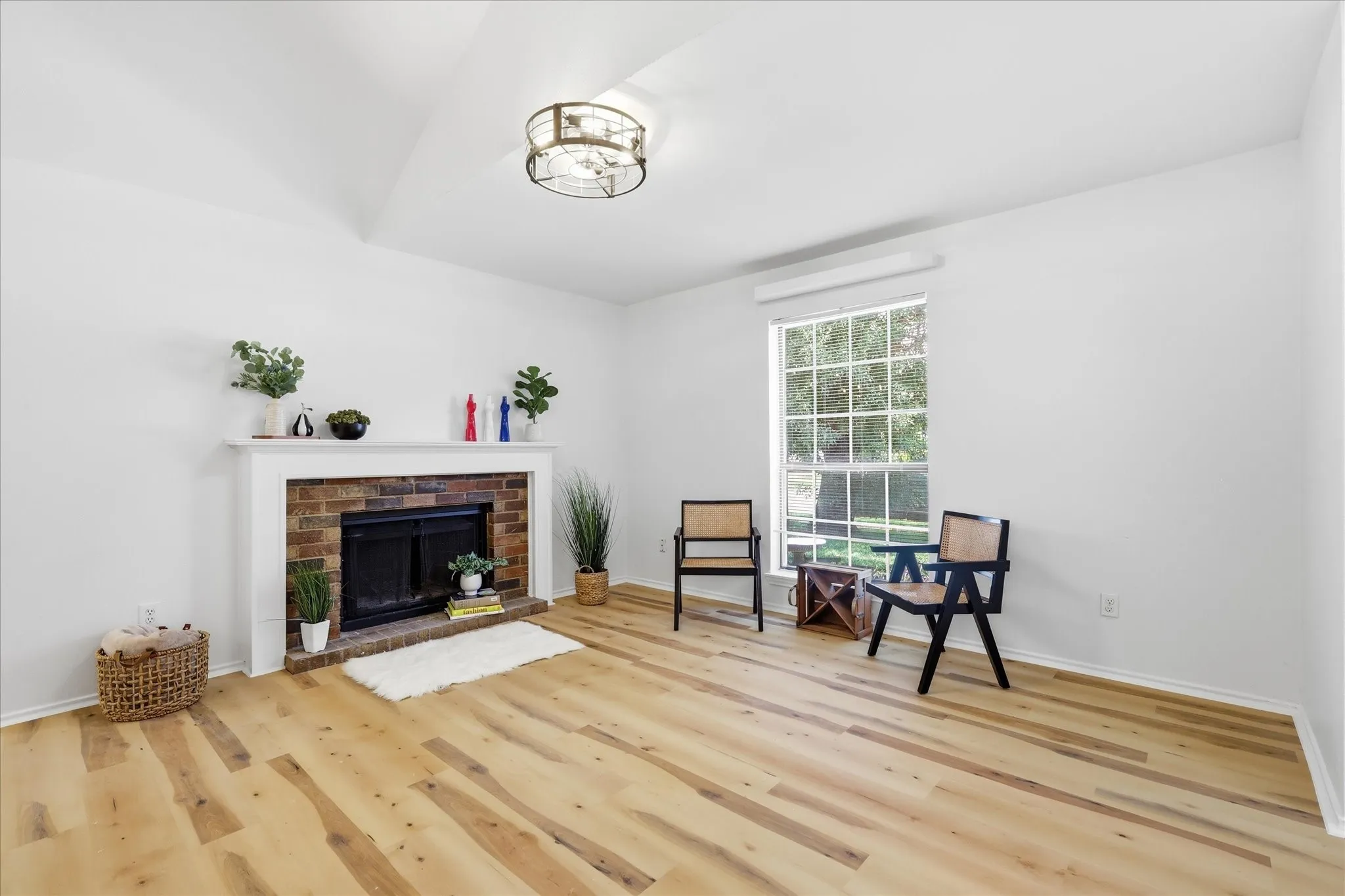 Vaulted ceiling with a flush-mount light fixture, brick fireplace with a white mantel, wood-finish flooring, and a large window with muntins