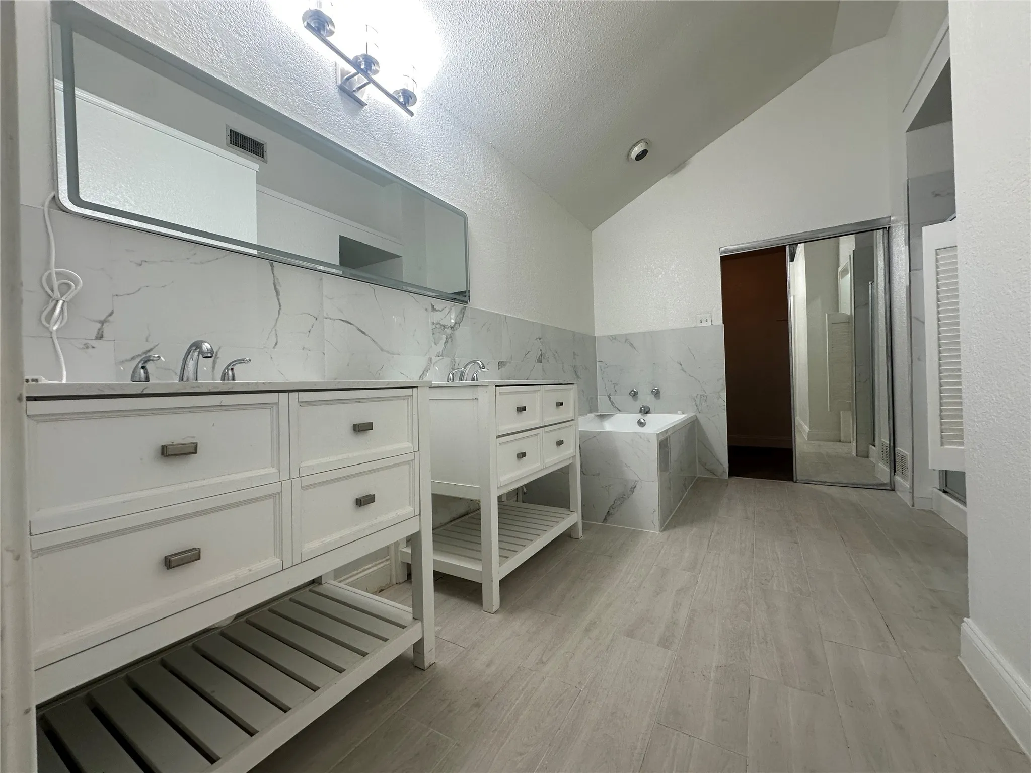 Dual vanity sinks with white cabinetry, a built-in tub with tile surround, wood-finish flooring, and a vaulted ceiling