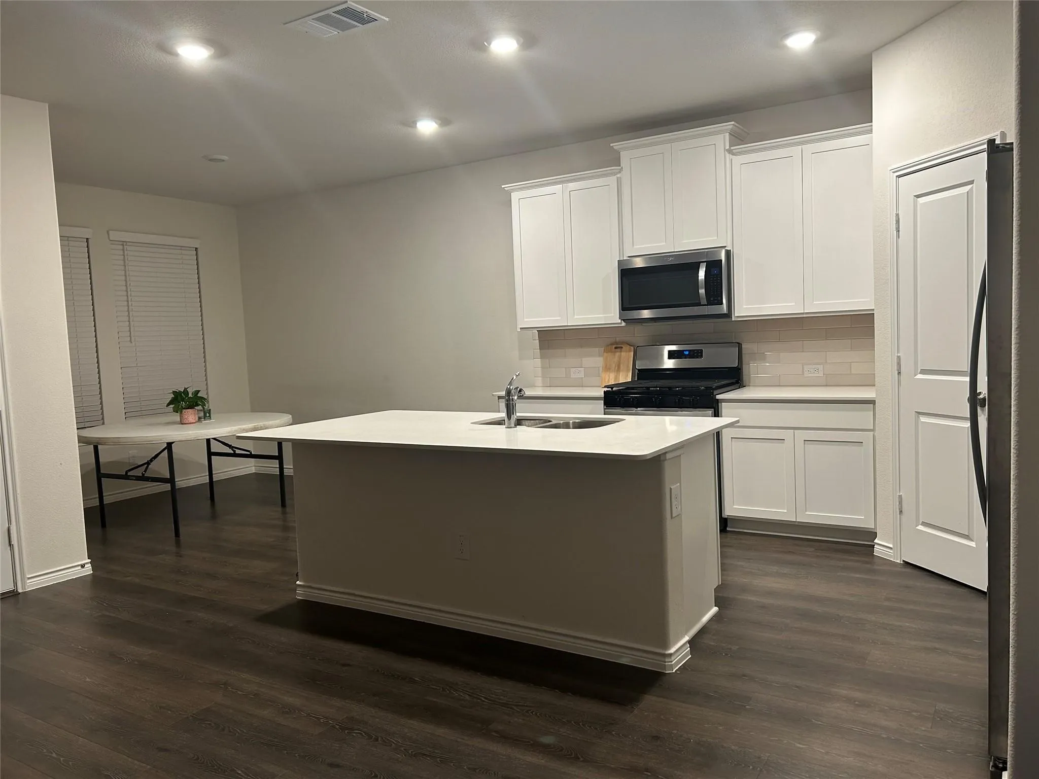 Kitchen featuring an island with an integrated sink, white cabinetry, stainless steel appliances, tile backsplash, and dark wood-finish flooring