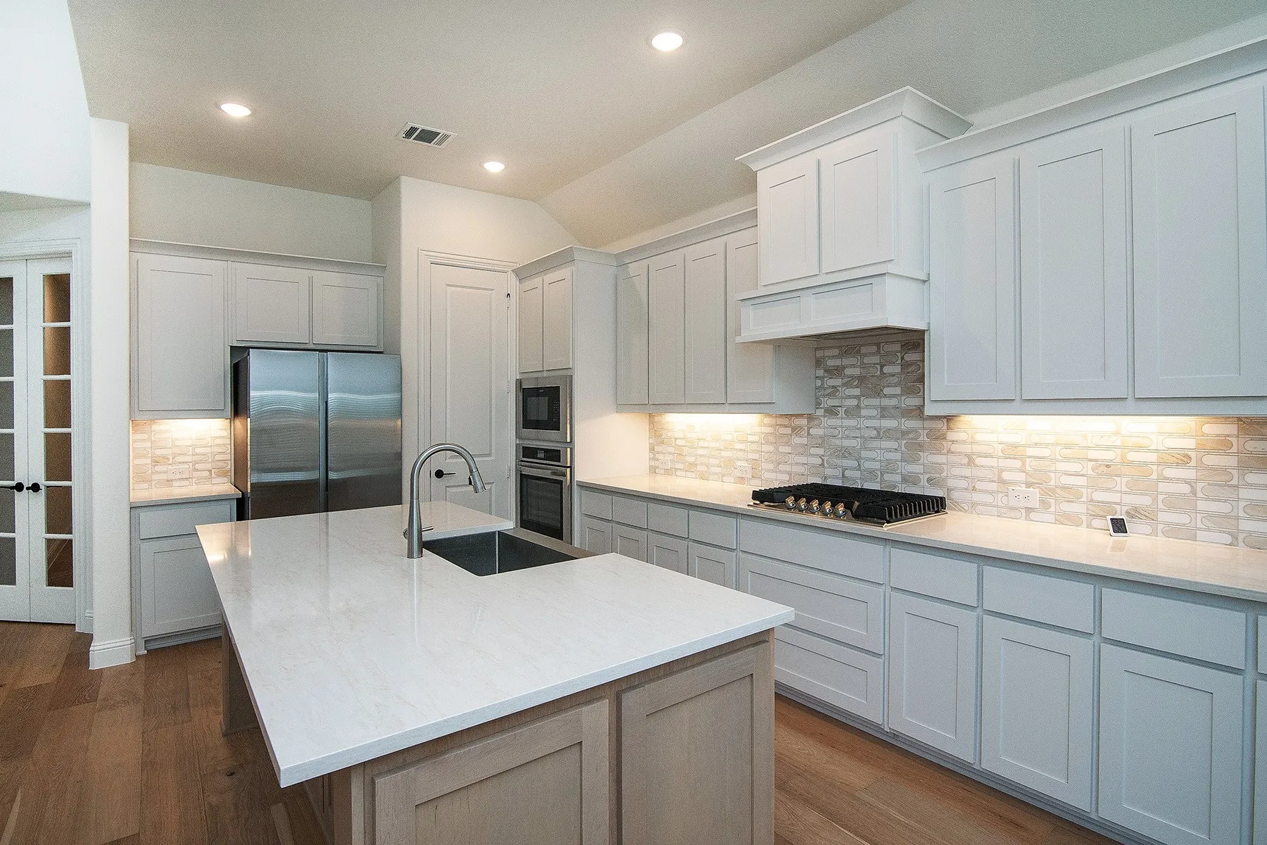 Kitchen featuring a large island with a surface-mounted sink, built-in stainless steel appliances, shaker-style cabinetry, a decorative range hood, and wood-finish flooring