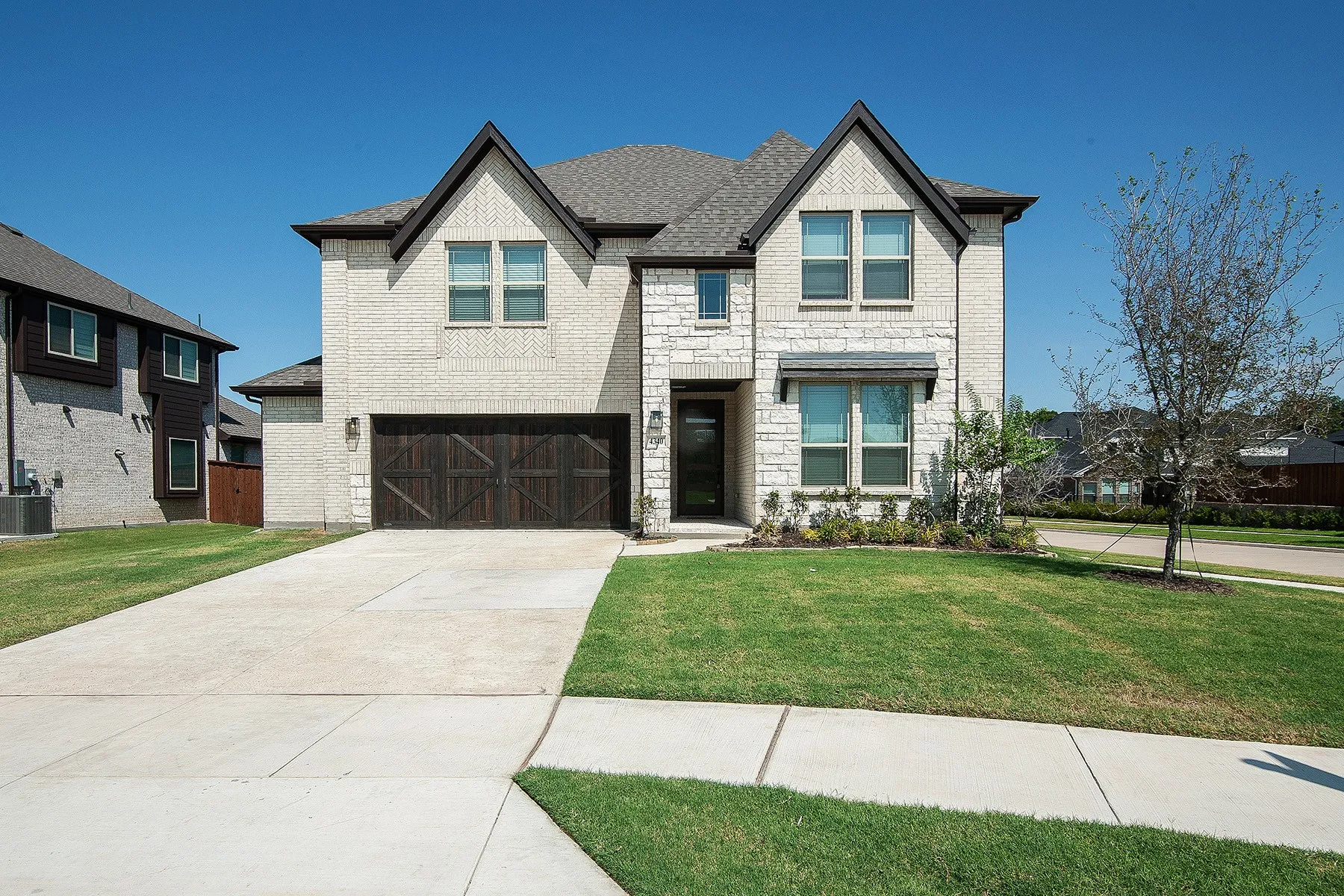 Two-story brick and stone exterior with dark trim accents, featuring a two-car garage with a wood-finish door, covered entryway, and concrete driveway