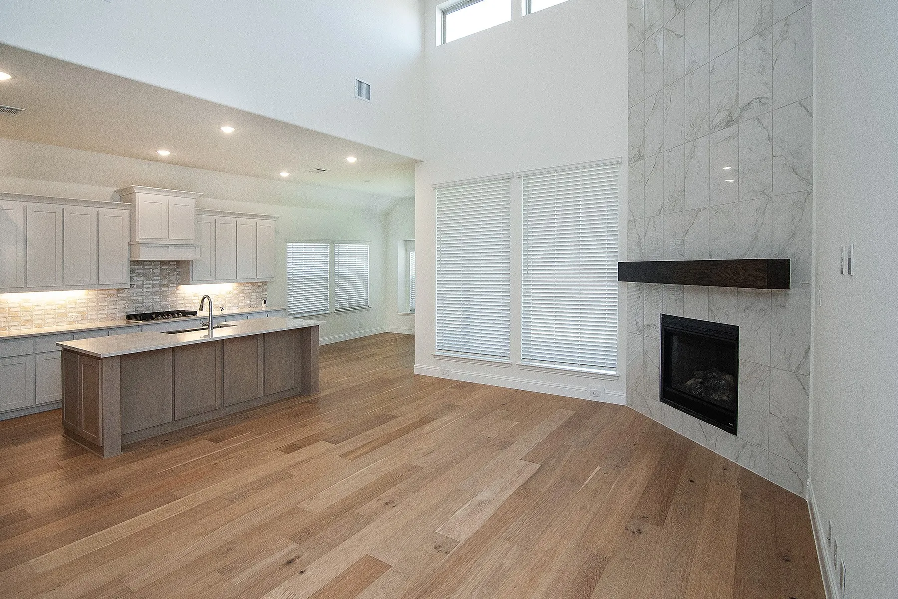 Wood-finish flooring, high ceilings, kitchen island with integrated sink, white cabinetry with under-cabinet lighting, and a fireplace with a stone-finish surround
