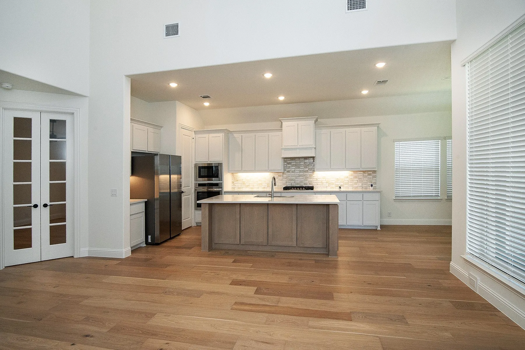 Open-concept kitchen and living area featuring wood-finish flooring, shaker-style cabinetry, a central island with a sink, stainless steel appliances, and French doors