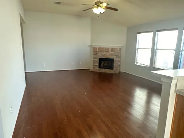 Spacious living area featuring wood-finish flooring, a brick fireplace with a stone mantel, a ceiling fan with integrated lighting, and multiple windows with blinds