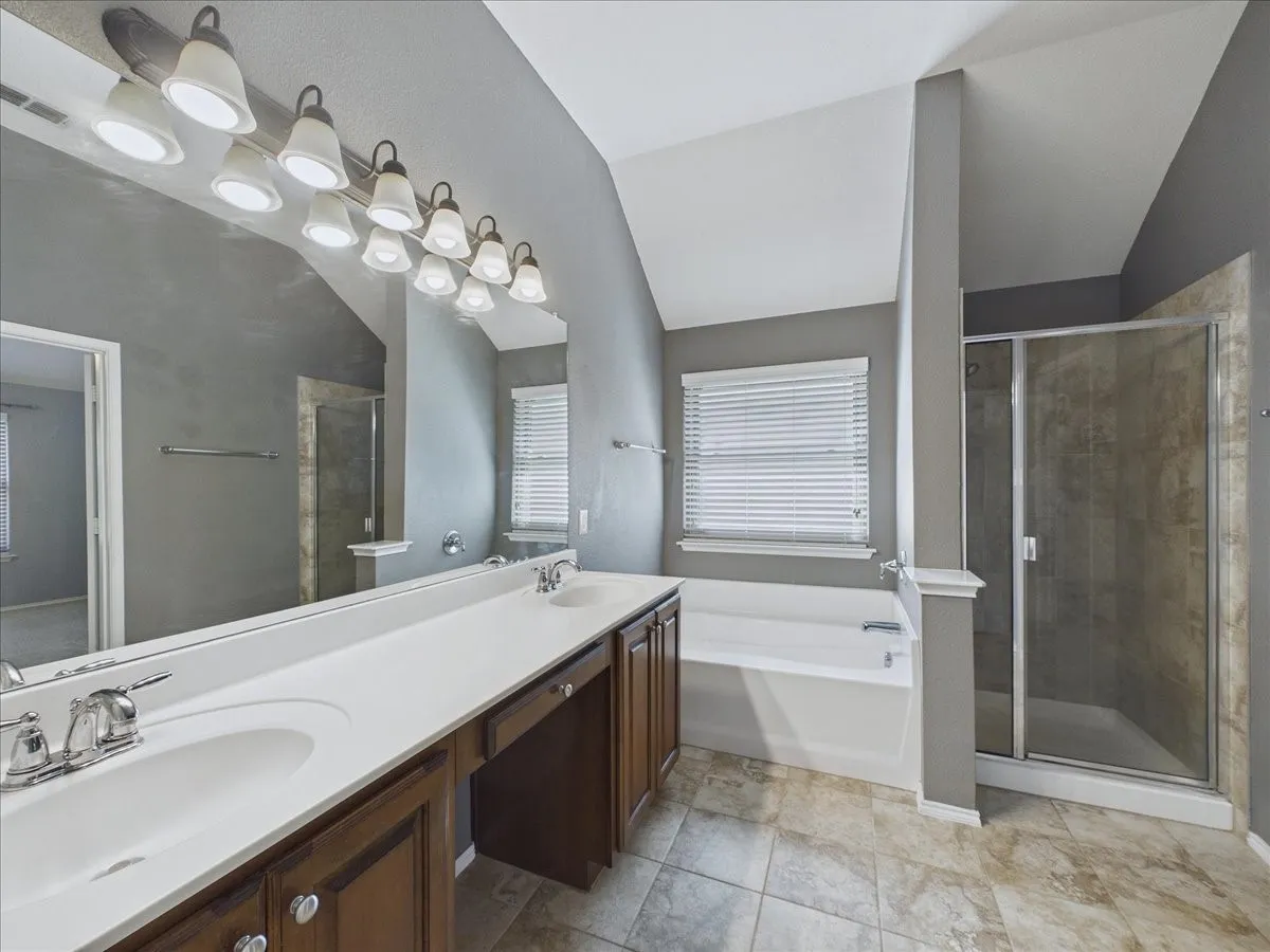 Dual vanity with integrated sinks, wood-finish cabinetry, built-in bathtub, glass-enclosed shower, and tiled flooring