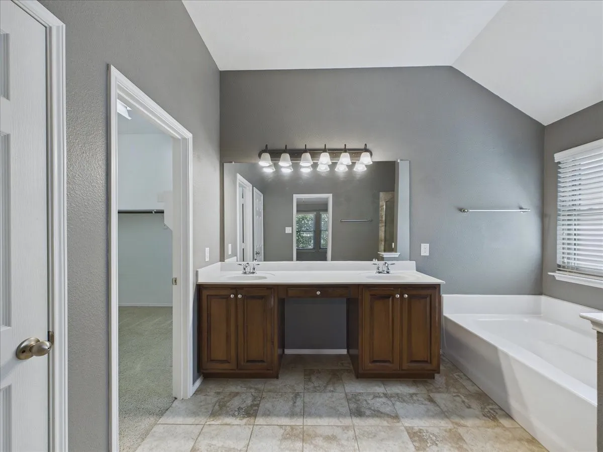 Spa-inspired primary bathroom featuring a dual-sink vanity with wood cabinetry, a large framed mirror with integrated lighting, a soaking tub, a walk-in shower with glass enclosure, tile flooring, and abundant natural light from multiple windows