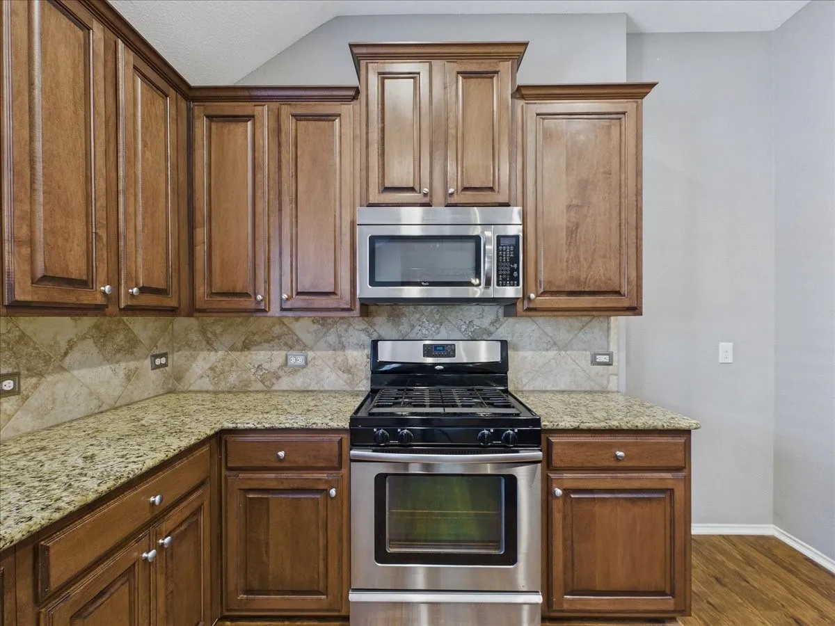 Wood-finish cabinetry with crown molding, granite-style countertops, stainless steel microwave, stainless steel gas range, and a tiled backsplash