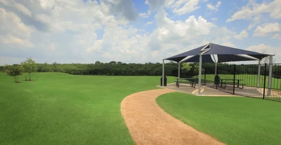 Expansive green space featuring a paved pathway, a shaded picnic area with metal fencing, and mature trees