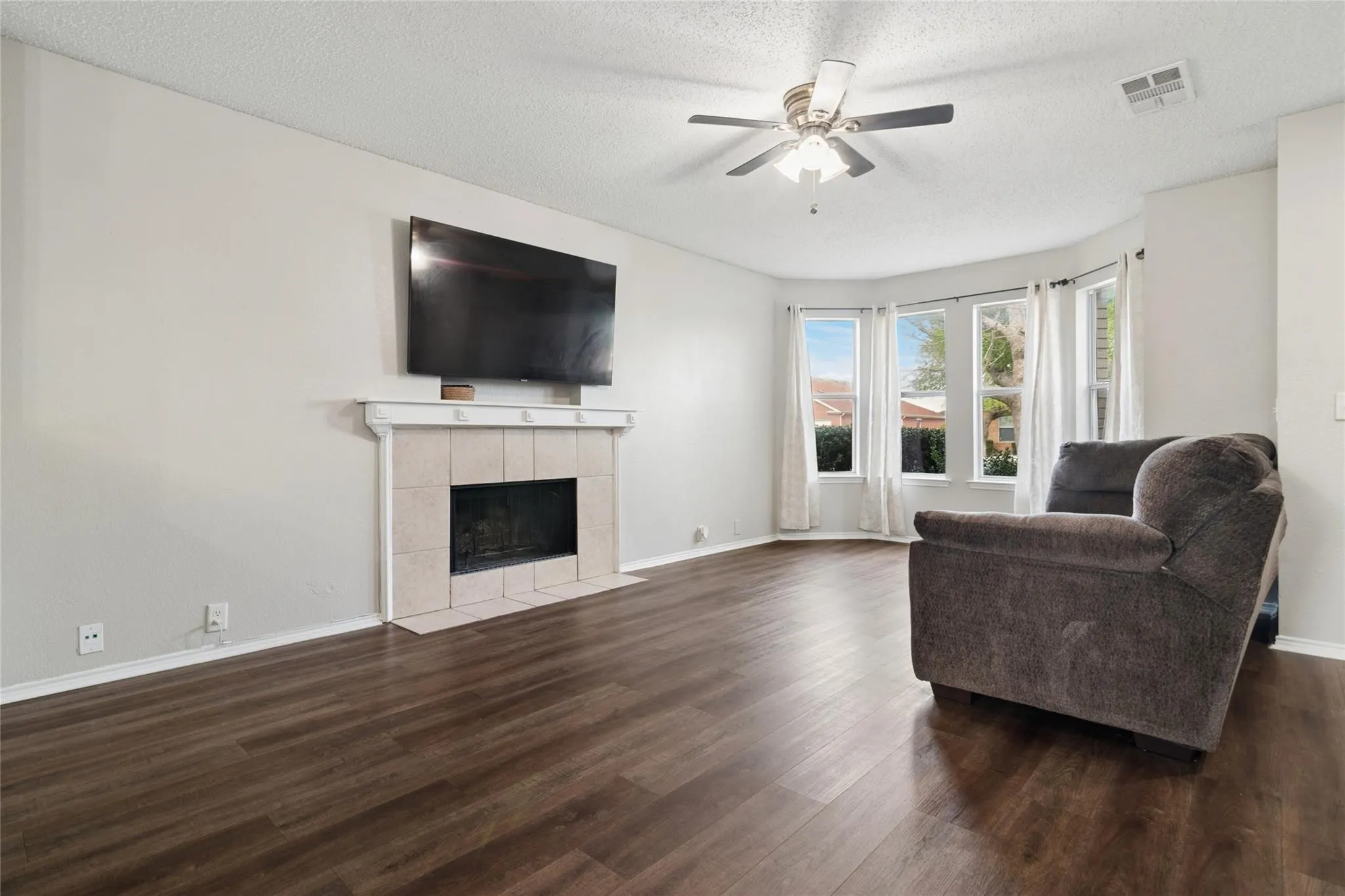 Bay window with multiple panes, wood-finish flooring, fireplace with tiled surround, ceiling fan, and light-toned walls