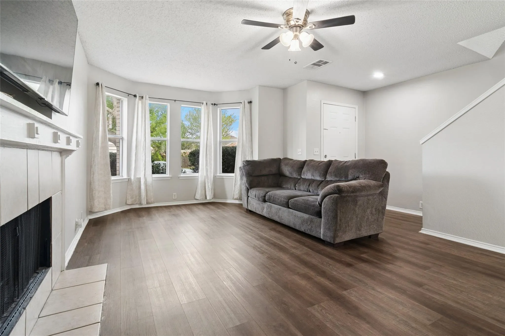 Spacious living area featuring wood-finish flooring, a fireplace with tile surround, a bay window, and a ceiling fan