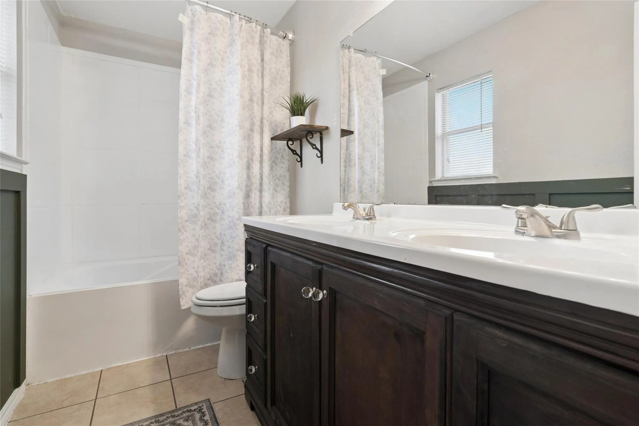 White bathtub with a shower, dark wood-finish double vanity, dual sinks with brushed nickel faucets, and tiled flooring