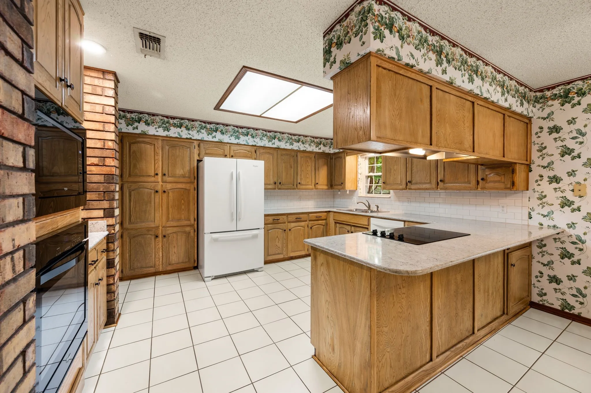 Kitchen featuring light-finish wood cabinetry, a stone-surfaced island with a cooktop, built-in wall ovens, exposed brick pillars, and ceramic tile flooring