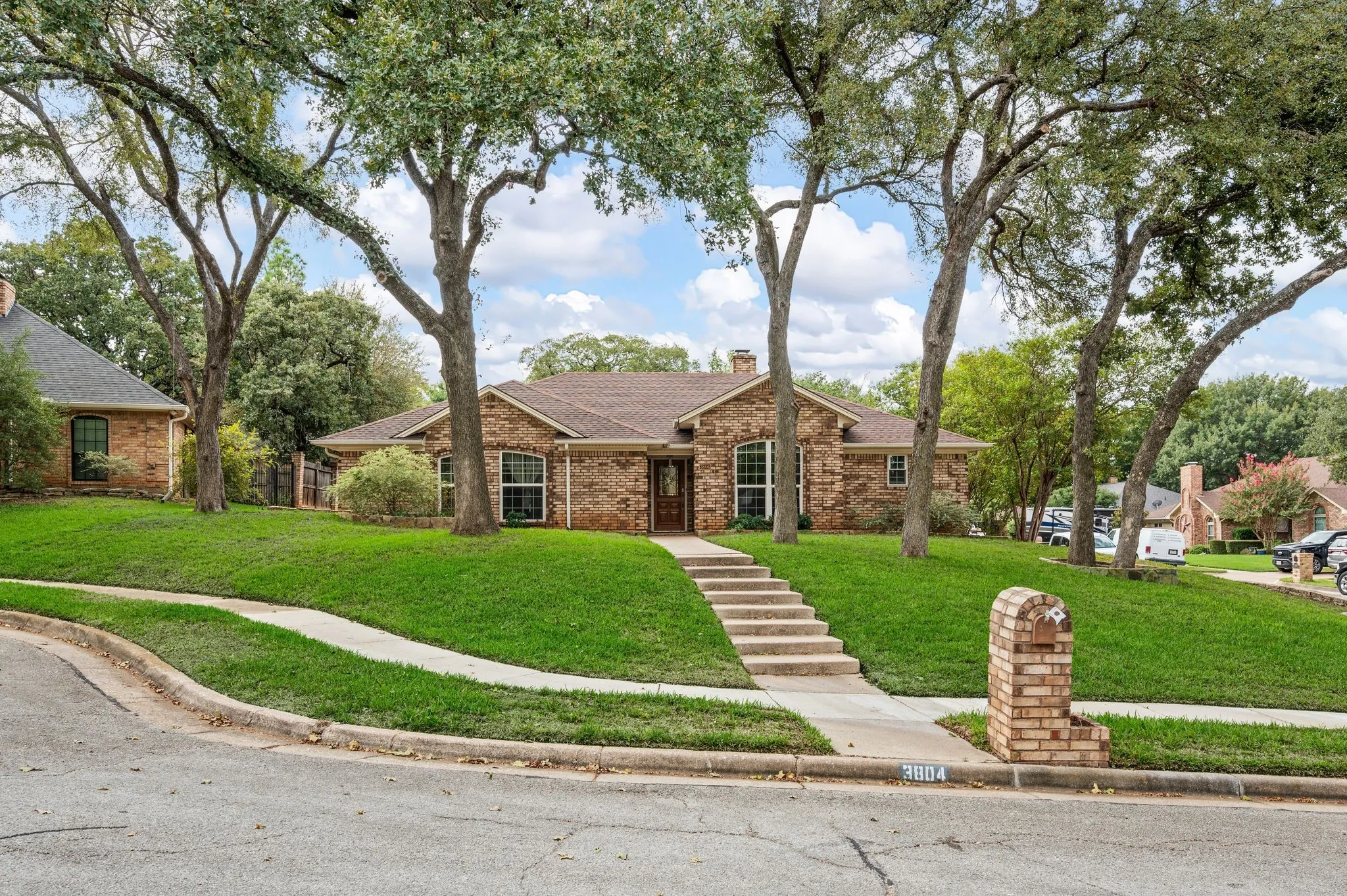 Single-story brick exterior featuring a multi-gabled roofline, arched window detailing, concrete steps, and a matching brick mailbox, all set on a landscaped lot with mature shade trees