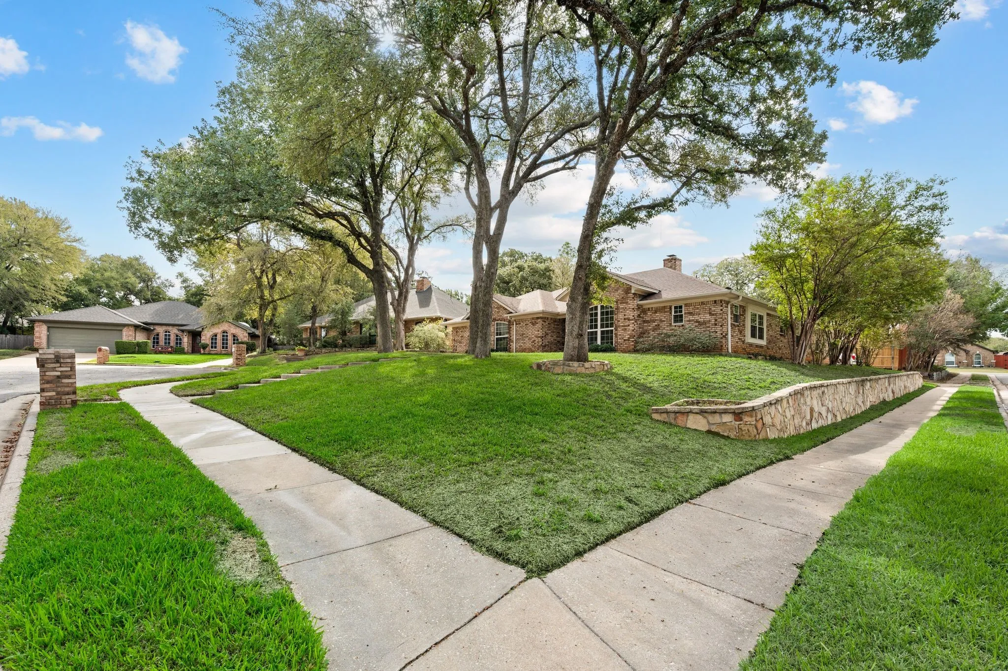 Brick exterior facade featuring a stone retaining wall, mature trees, and a concrete sidewalk