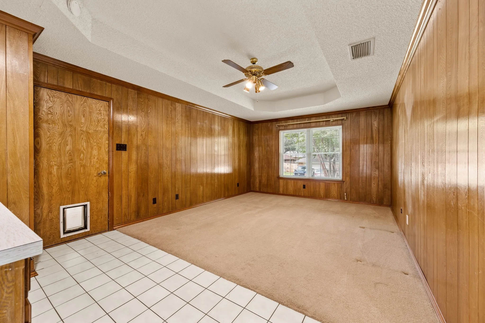 Tray ceiling with a central multi-light ceiling fan, extensive wood paneling, tile flooring, and a window with exterior scenery