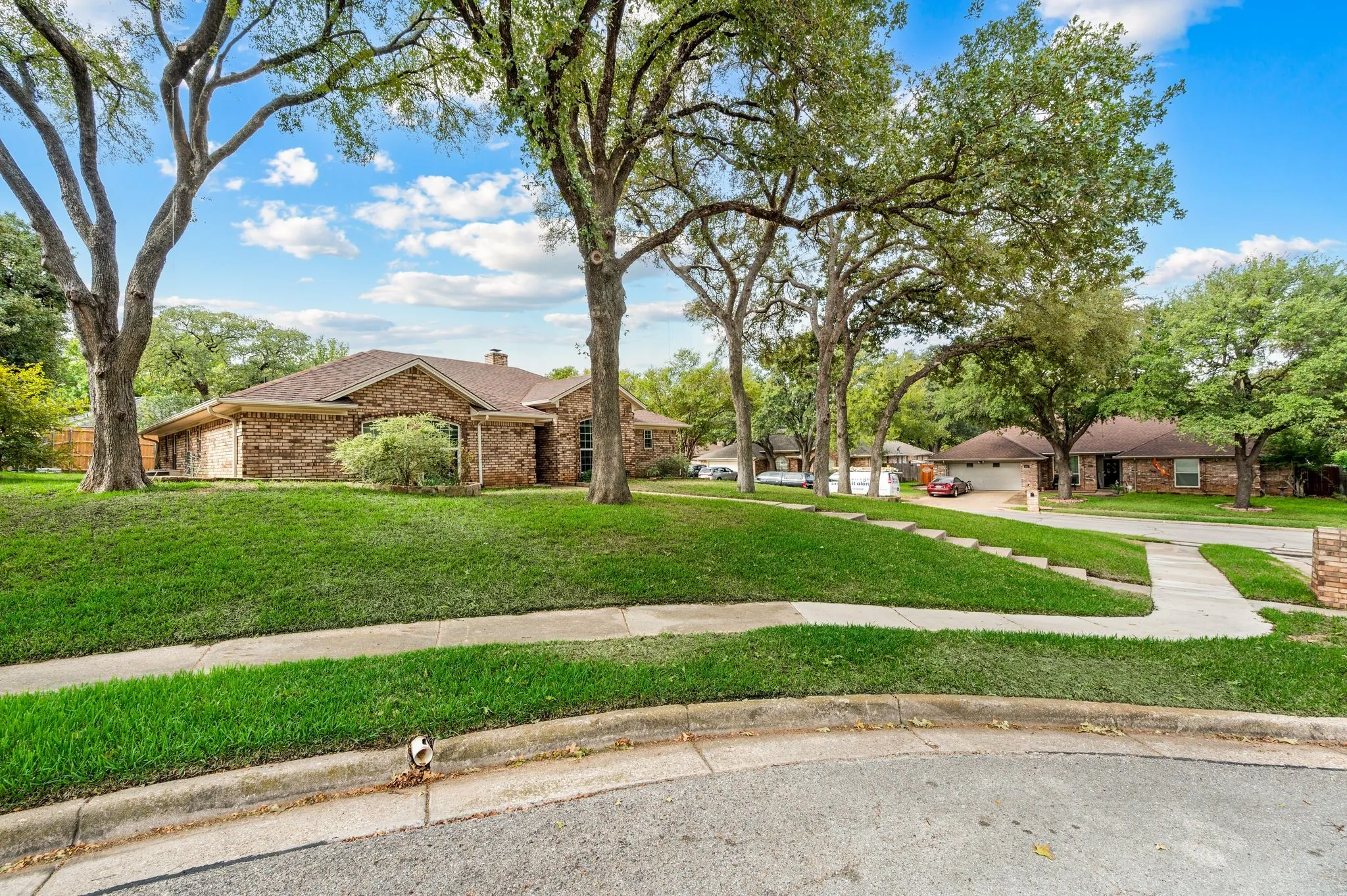 Brick exterior residence featuring a hip roof, concrete sidewalk, mature shade trees, and a landscaped lawn