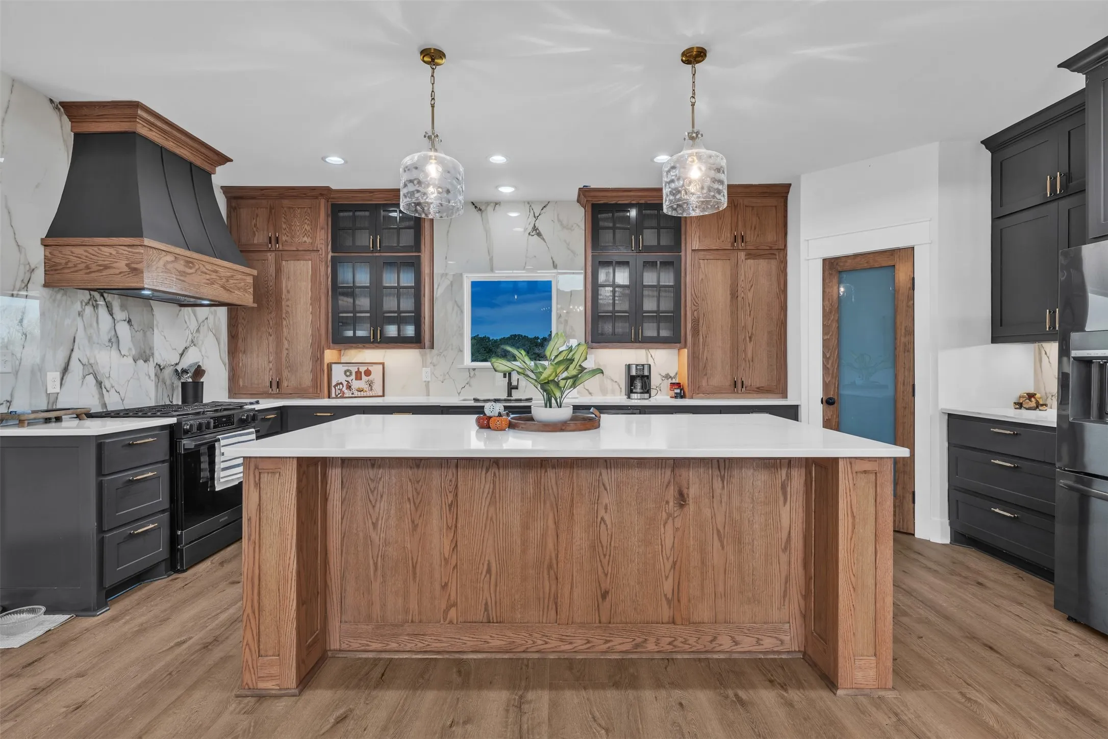 Wood-finish flooring, a white countertop island, custom wood-tone cabinetry, a black range hood, and pendant lighting