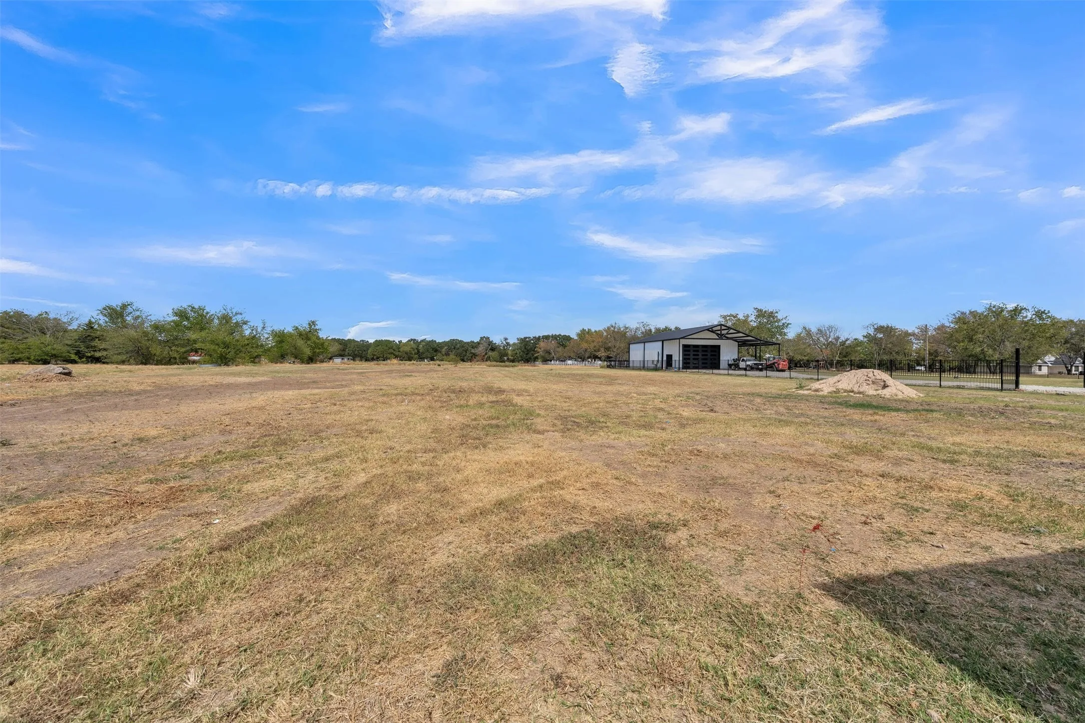 Expansive land parcel featuring a metal-sided structure with an attached covered area, partially fenced perimeter, and distant tree line