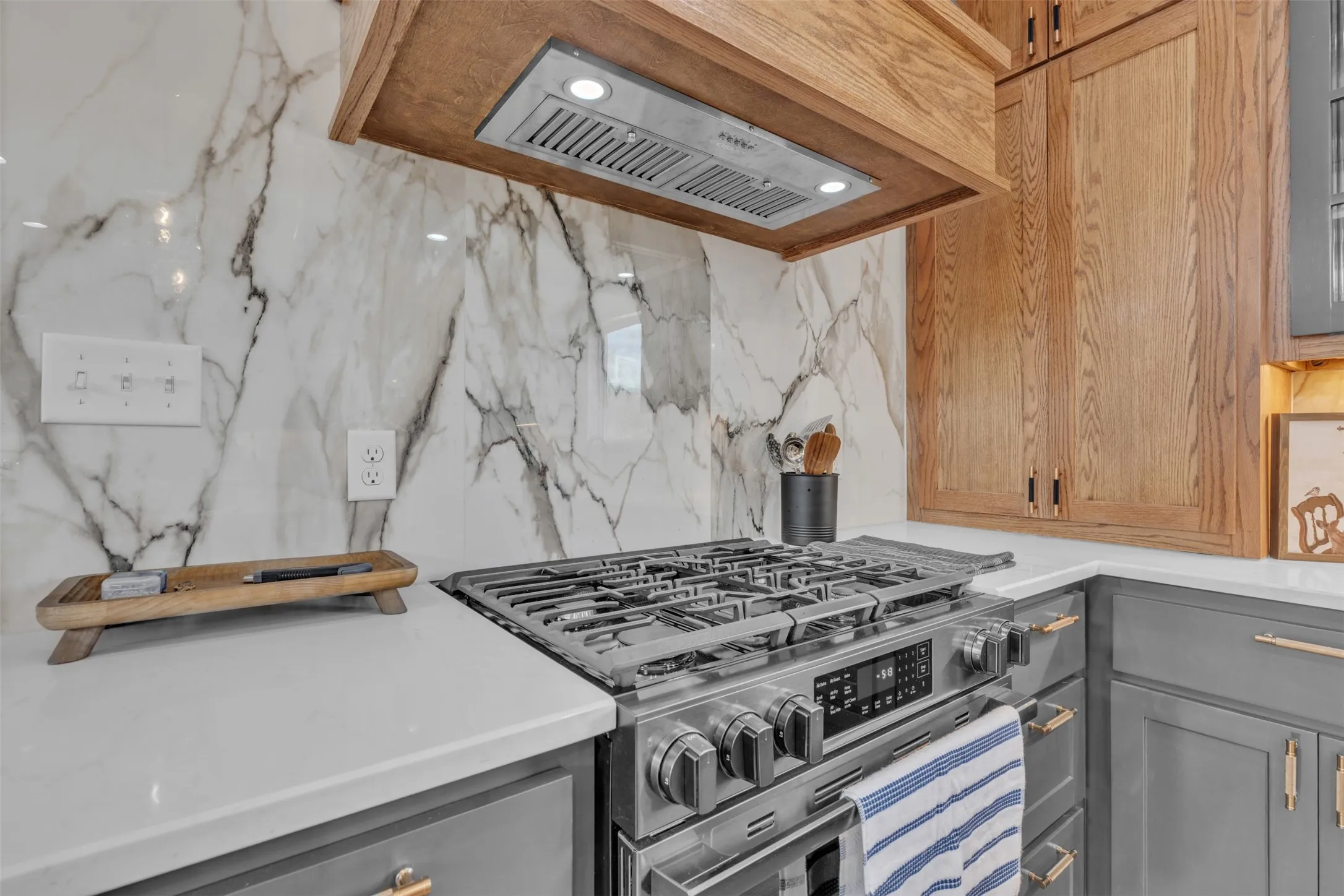 Stainless steel gas range, wood-finish range hood, white countertop, marble-look backsplash, and grey lower cabinetry