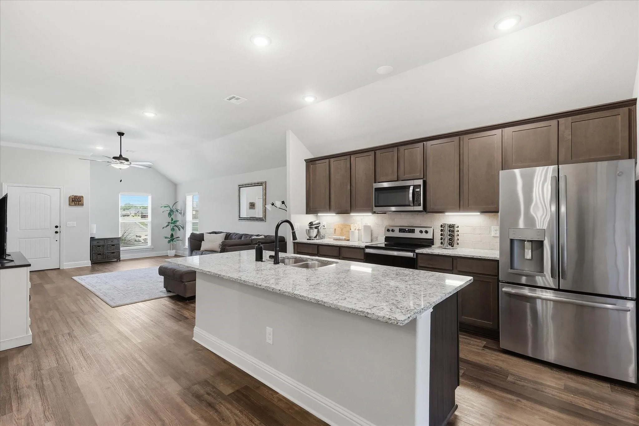 Spacious kitchen island with light-toned countertop, stainless steel appliances, dark wood-finish cabinetry, wood-finish flooring, and vaulted ceiling